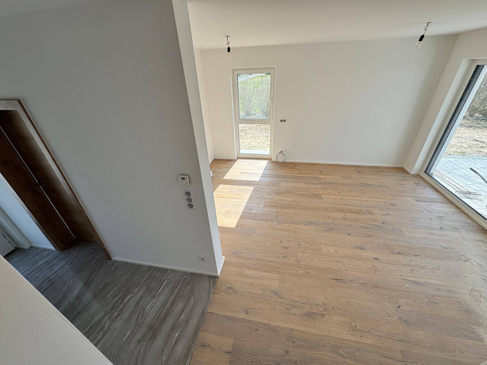 Geräumiger Wohnbereich mit hellem Holzboden und großen Fenstern, die viel Tageslicht hereinlassen.
