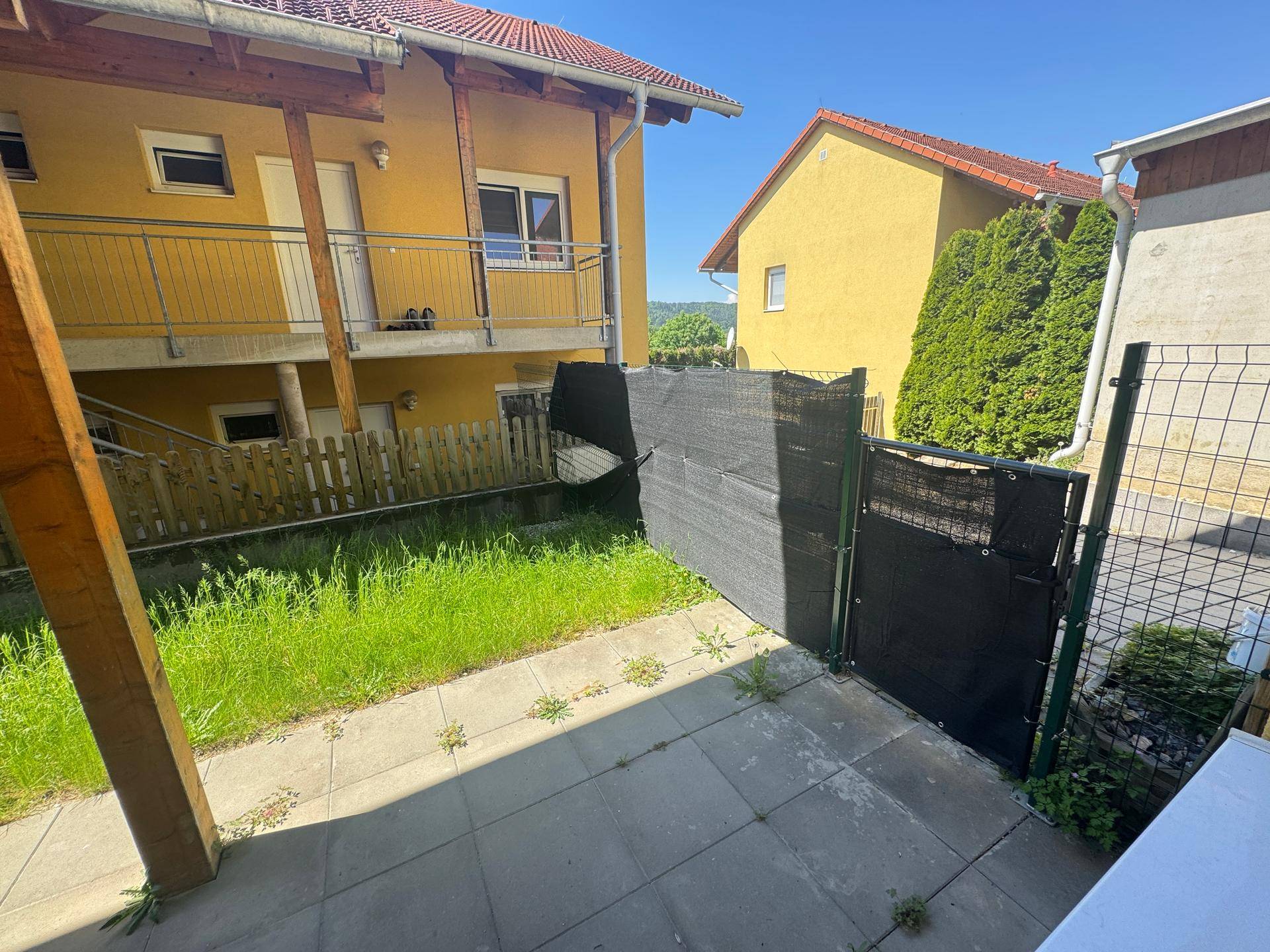 Gepflasterte Terrasse mit Rasenfläche und einem schwarzen Sichtschutzzaun, der zum Garten führt.