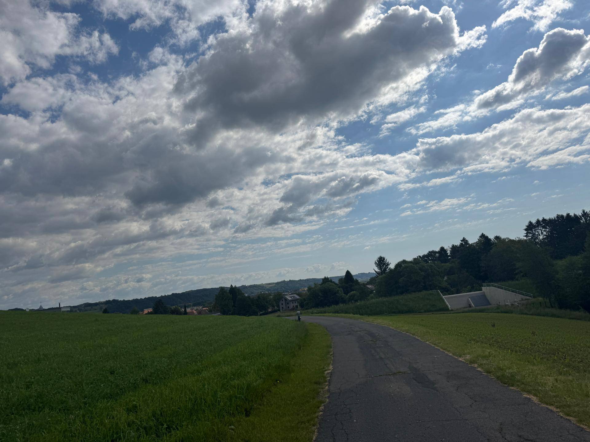 Landstraße schlängelt sich durch eine weite grüne Landschaft unter einem dynamischen Wolkenhimmel.