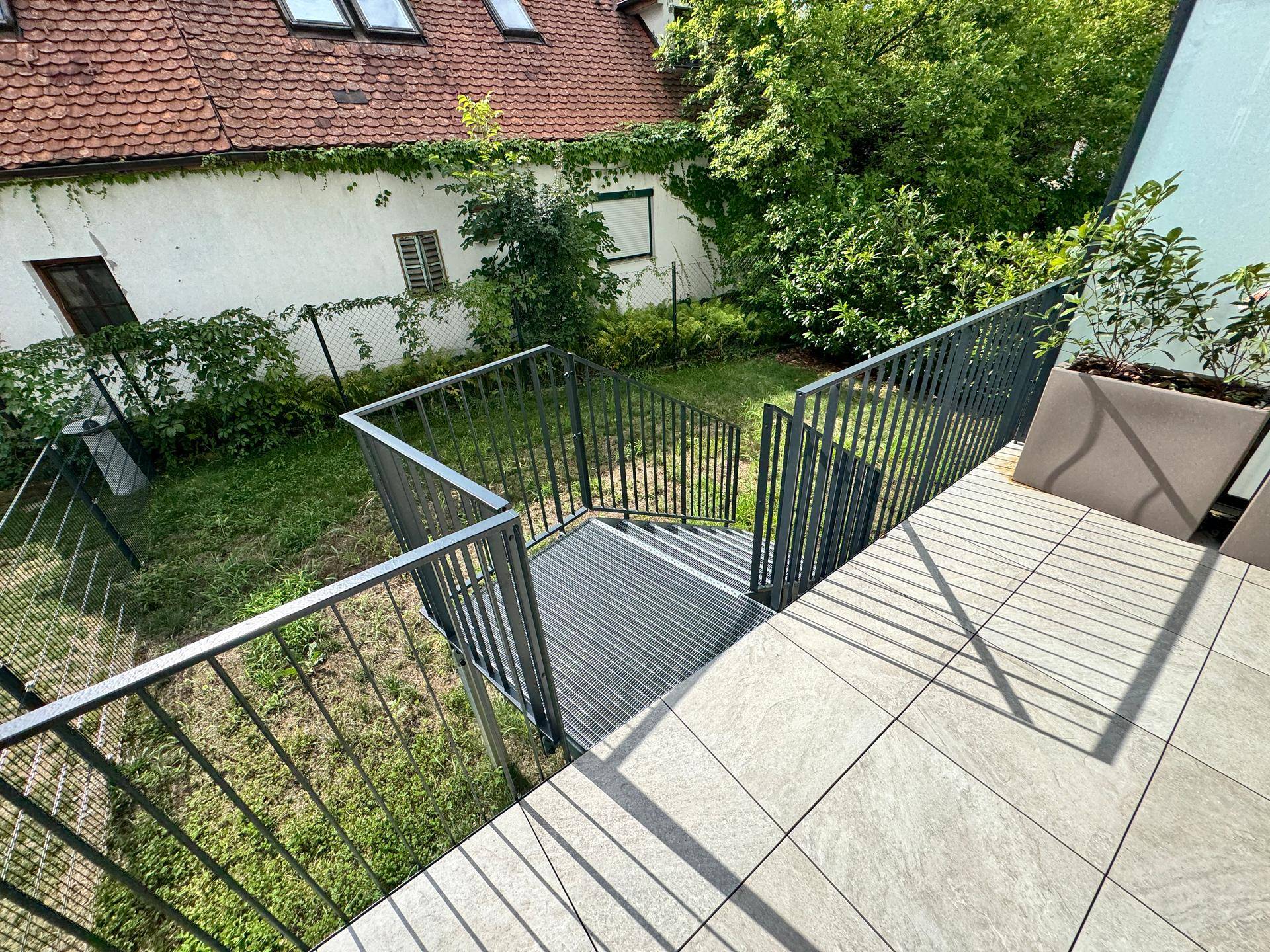 Blick von einer Terrasse auf einen grünen Gartenbereich und ein angrenzendes älteres Gebäude.