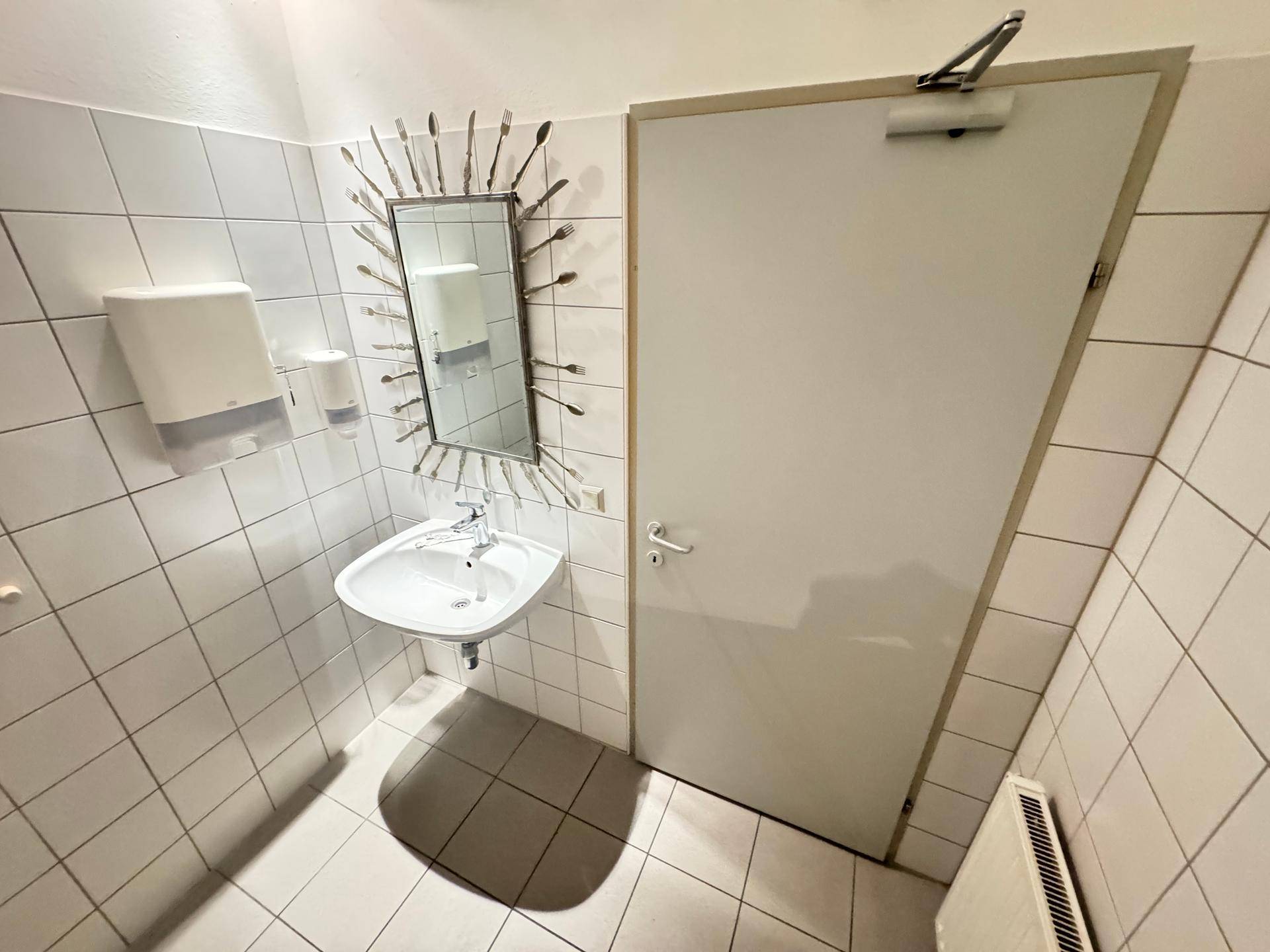 Einfaches WC mit weiß gefliesten Wänden, einem Waschbecken und einem Spiegel.