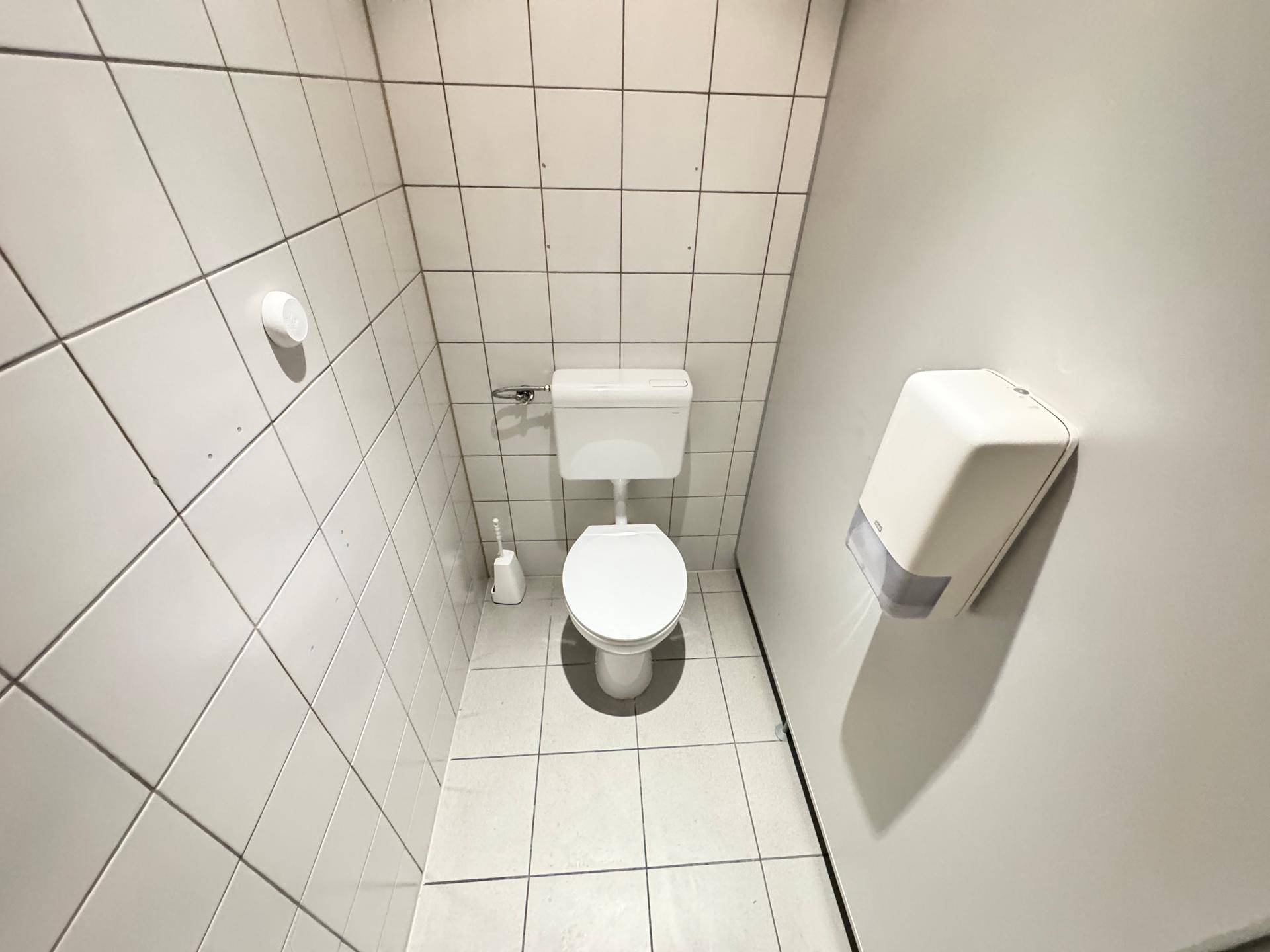 Sauberes WC mit weiß gefliesten Wänden und Boden, ausgestattet mit einer Toilette und Spender.