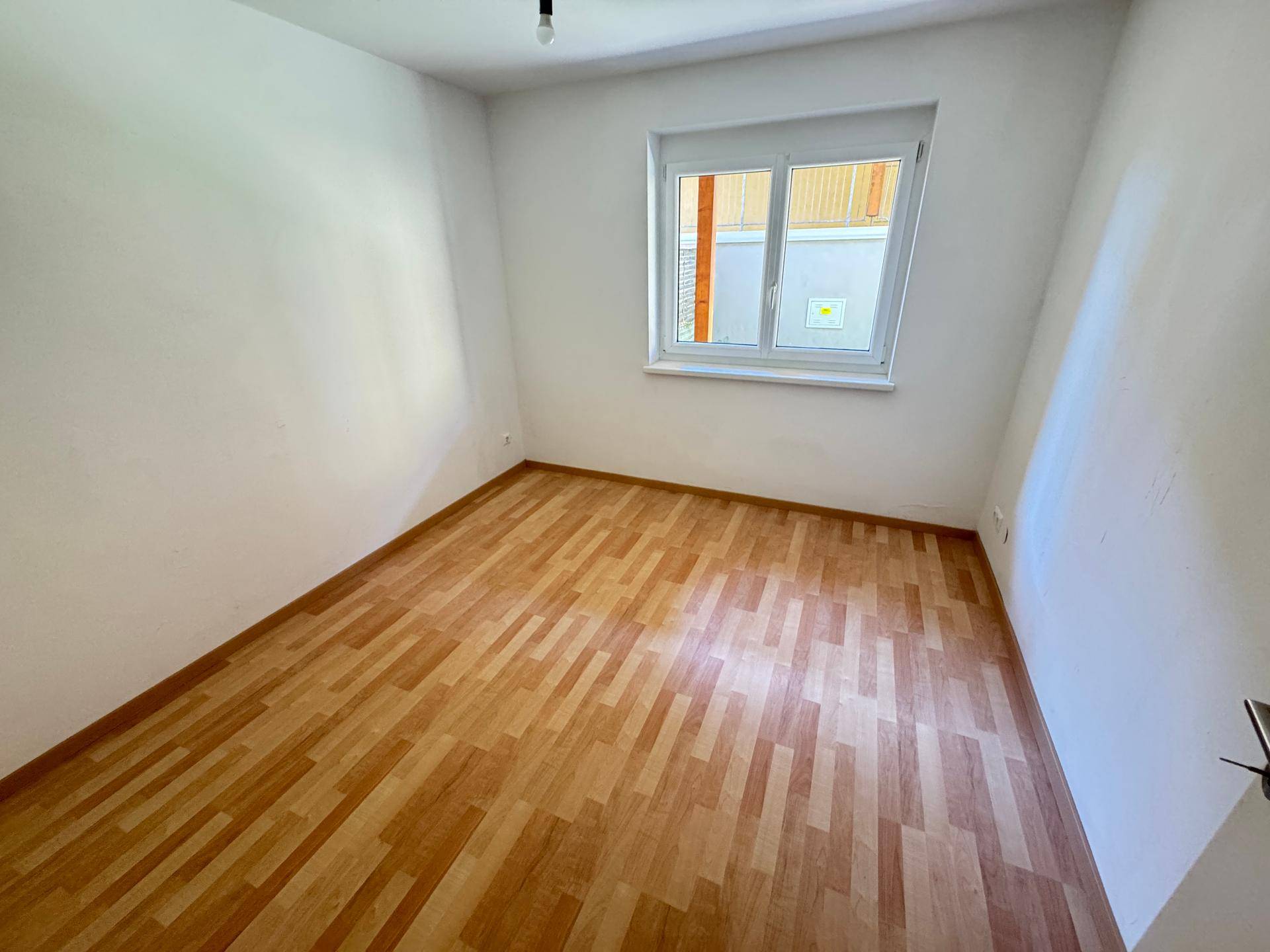 Leeres Zimmer mit Laminatboden und einem Fenster, das Tageslicht in den Raum bringt.