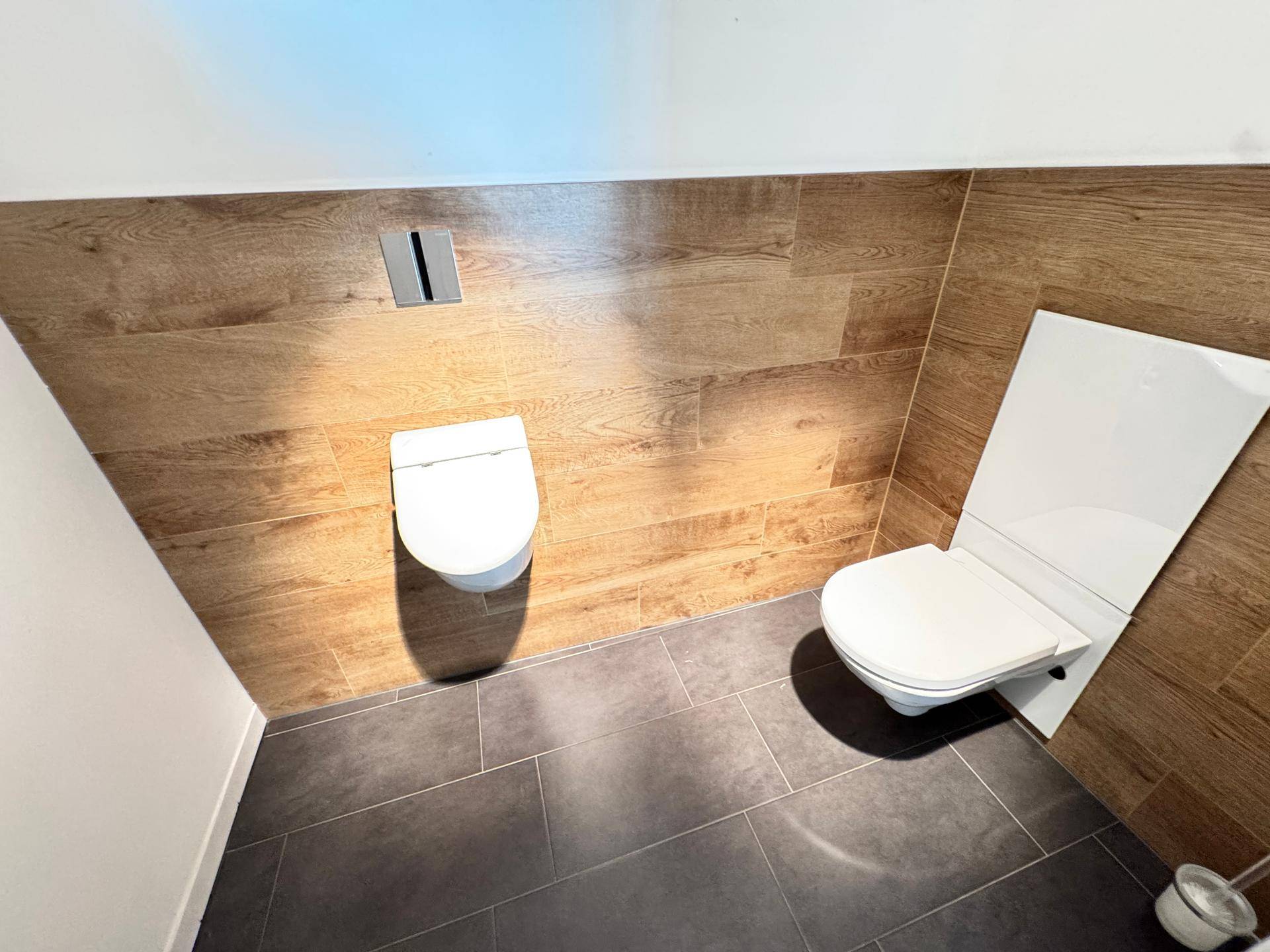 Modernes WC mit zwei wandhängenden Toiletten, dunklen Fliesen und Holzoptik-Wandverkleidung.