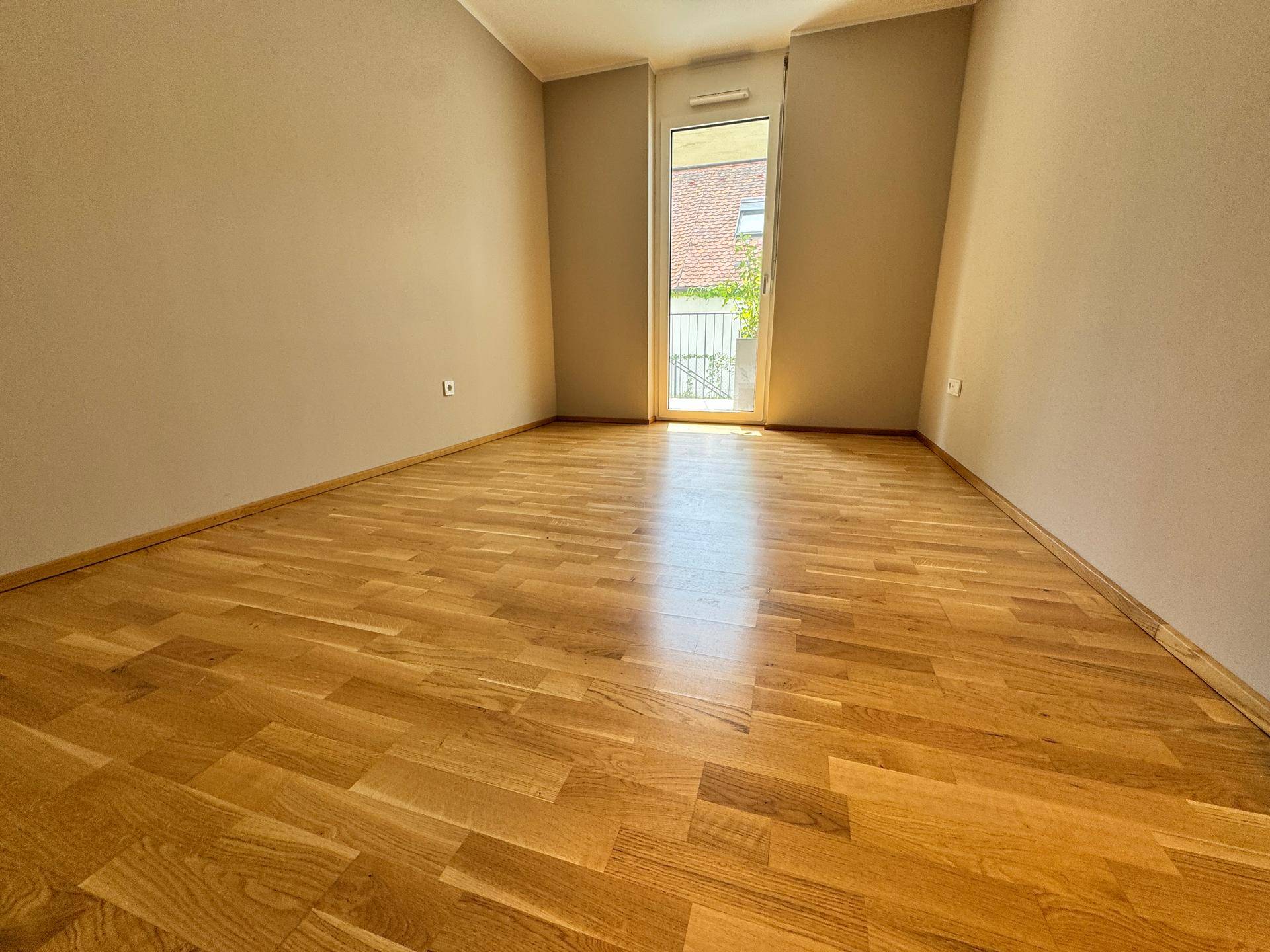 Leeres Schlafzimmer mit Holzboden und Zugang zu einem französischen Balkon, bietet viel Tageslicht.