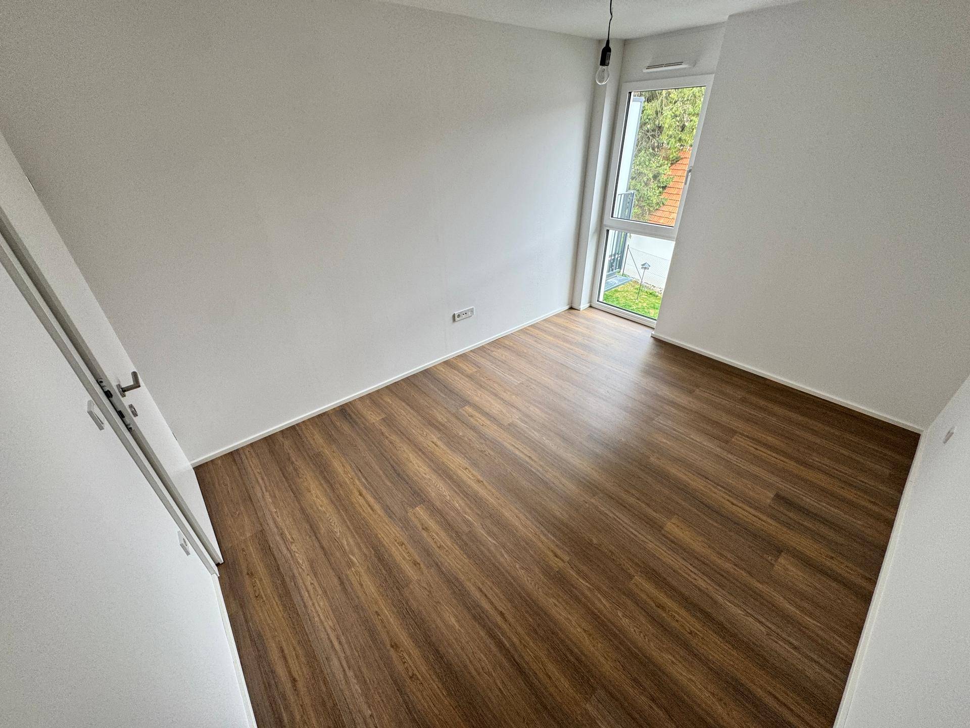 Leeres Schlafzimmer mit Laminatboden und einem hohen Fenster, das viel Licht hereinlässt.