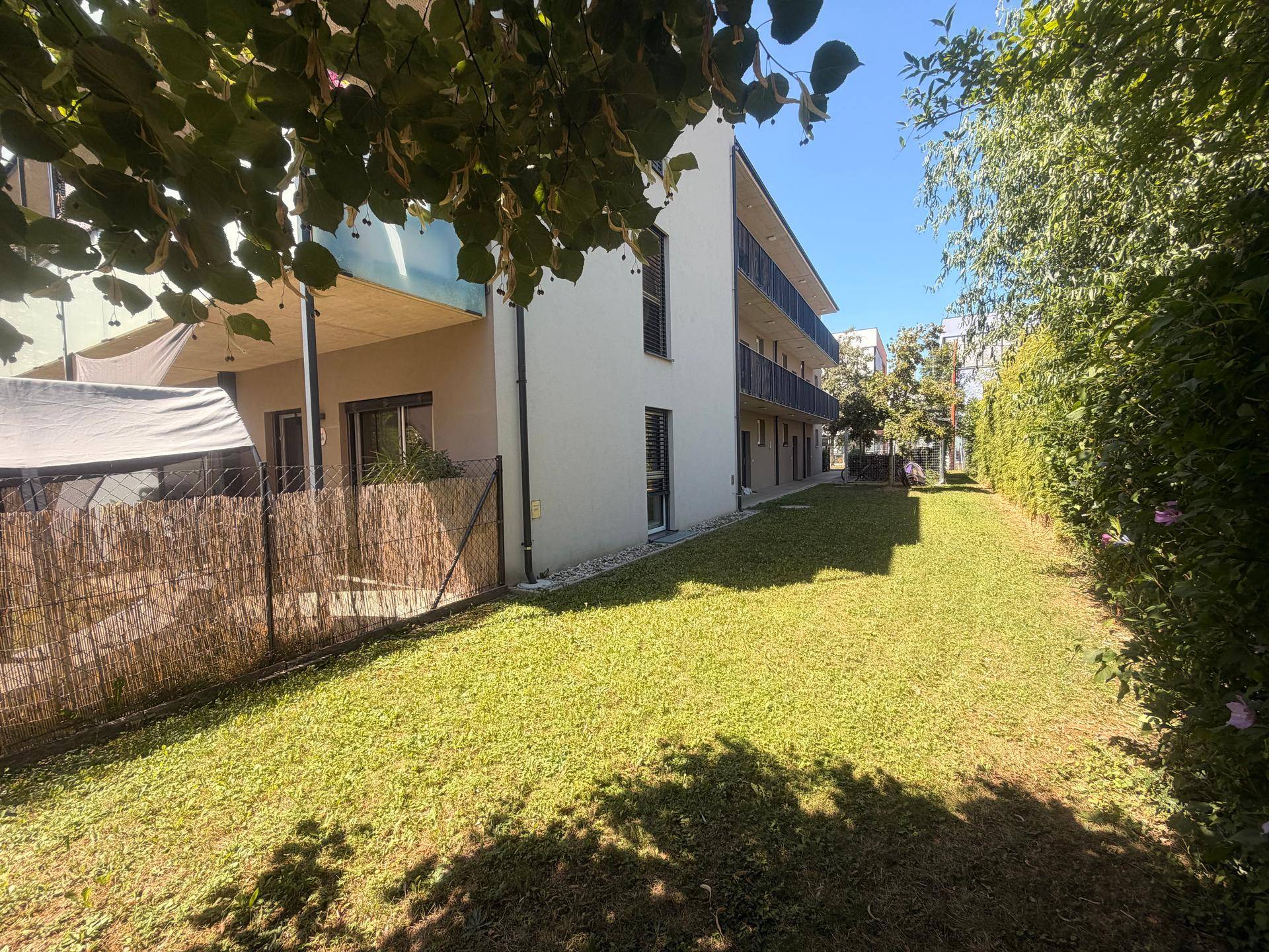 Weitläufiger Gartenbereich mit Rasen und Bäumen, angrenzend an die Gebäudeseite mit Balkonen.