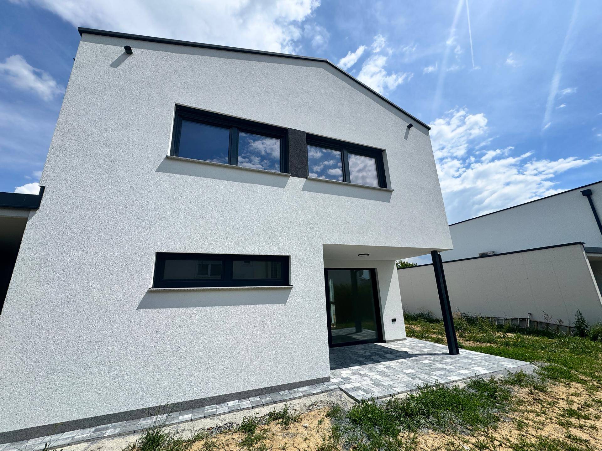 Neubau mit moderner Architektur, weißer Fassade, großen Fenstern und einer gepflasterten Terrasse.