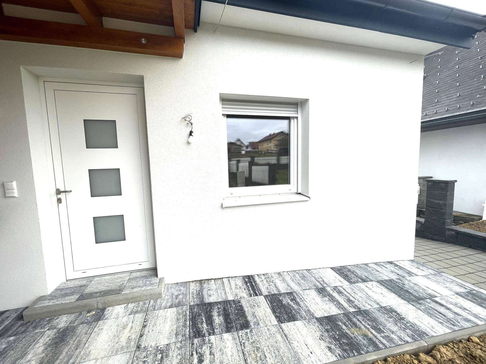 Moderner Eingangsbereich mit weißer Tür, Fenster und gefliester Terrasse, einladend gestaltet.