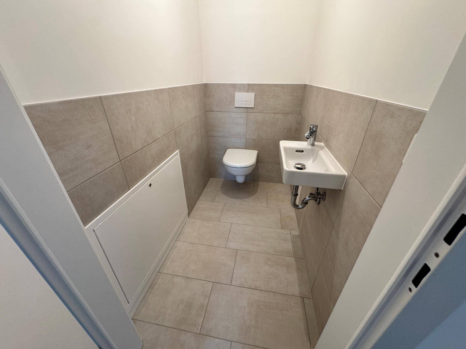 Modernes Gäste-WC mit Wand-WC, kleinem Waschbecken und hellen Fliesen.