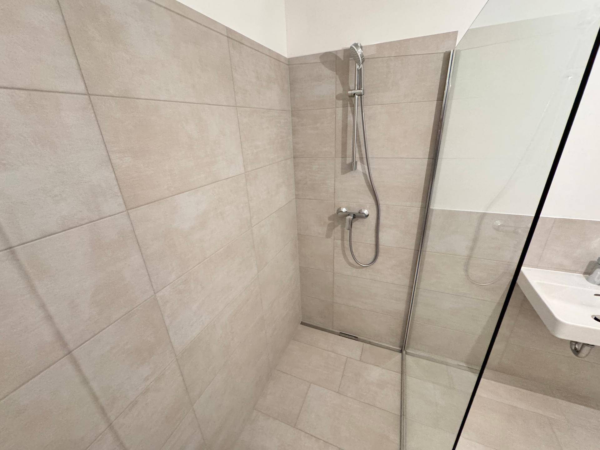 Detailansicht einer modernen, begehbaren Dusche mit Glaswand und beigefarbenen Fliesen.