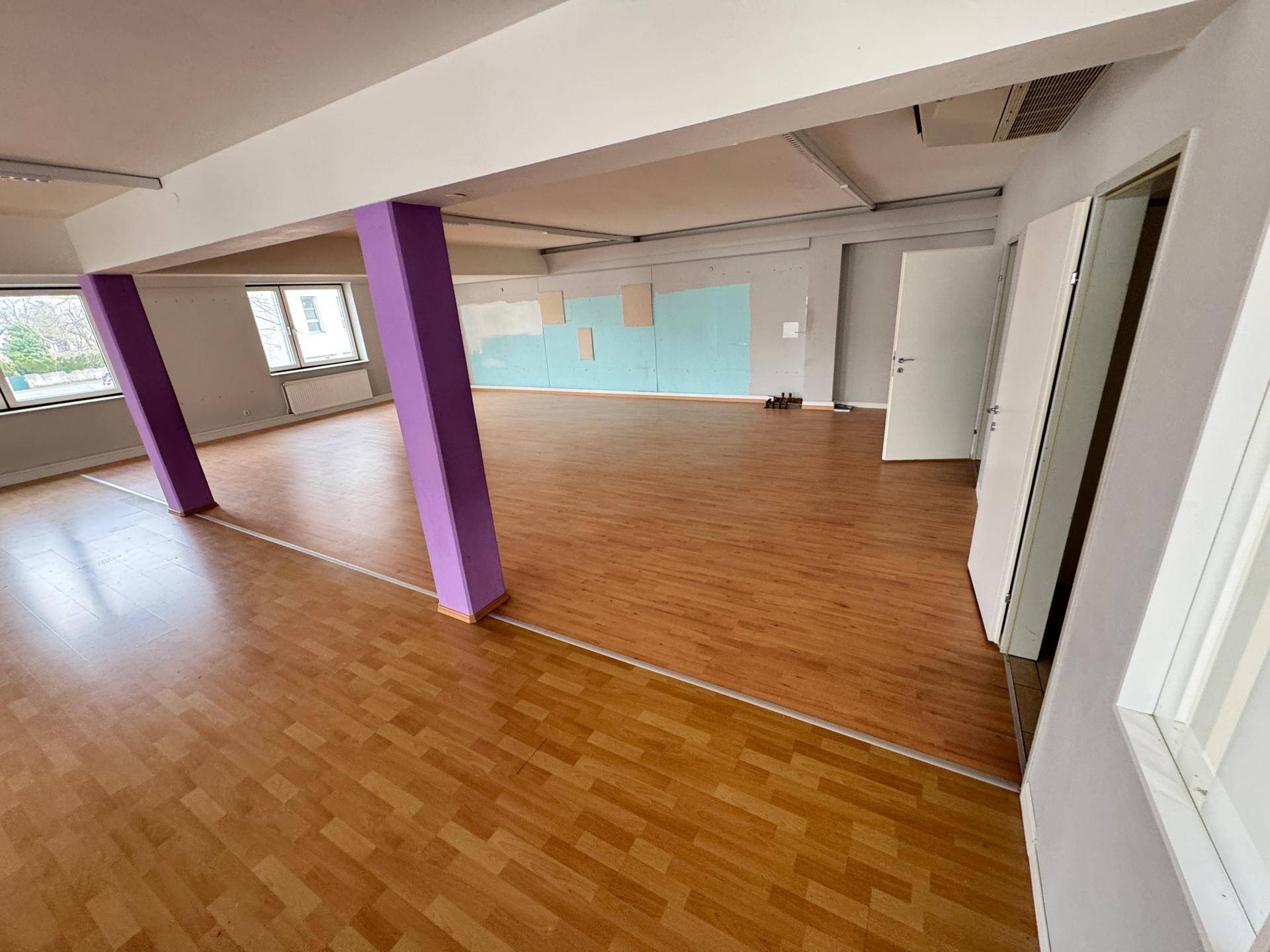 Großer offener Raum mit hellem Laminatboden und markanten lila Stützpfeilern.