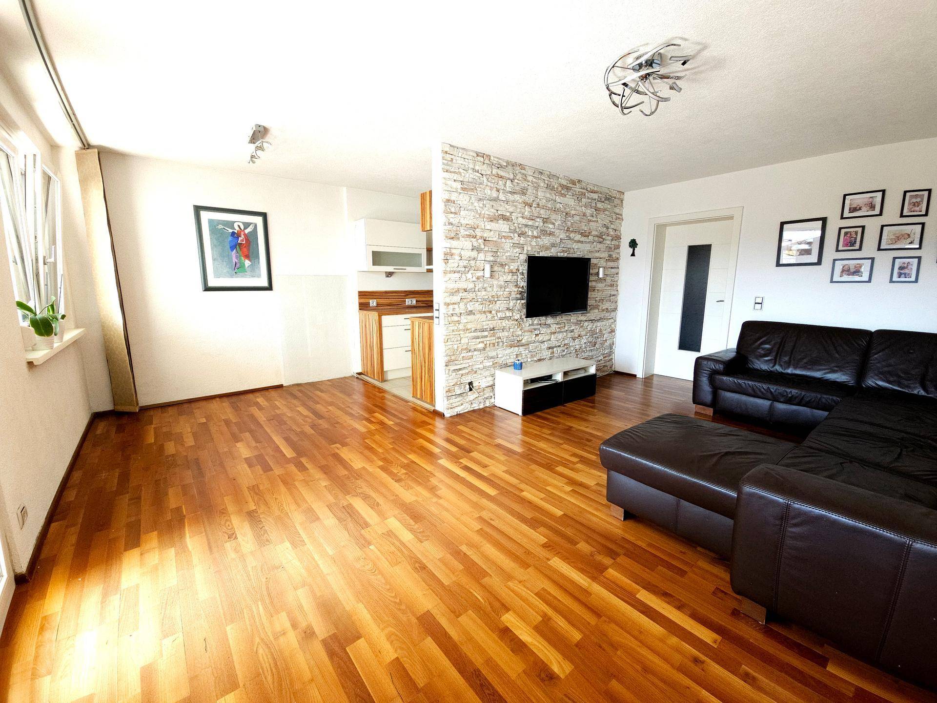 Geräumiges Wohnzimmer mit Holzboden, Steinwand und Blick in die angrenzende Küche.