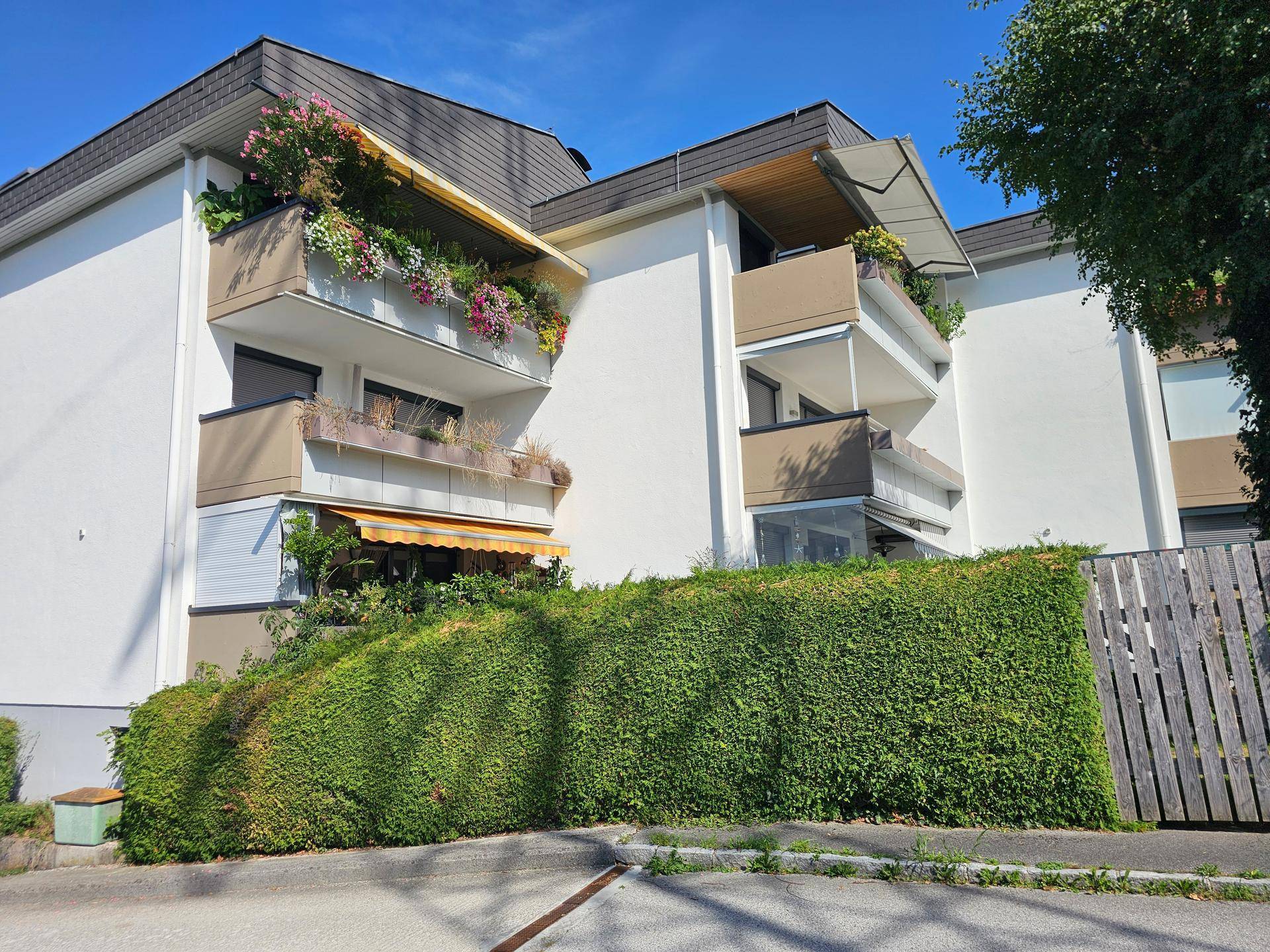 Mehrfamilienhaus mit Balkonen, Markisen und gepflegter Hecke im Vordergrund.