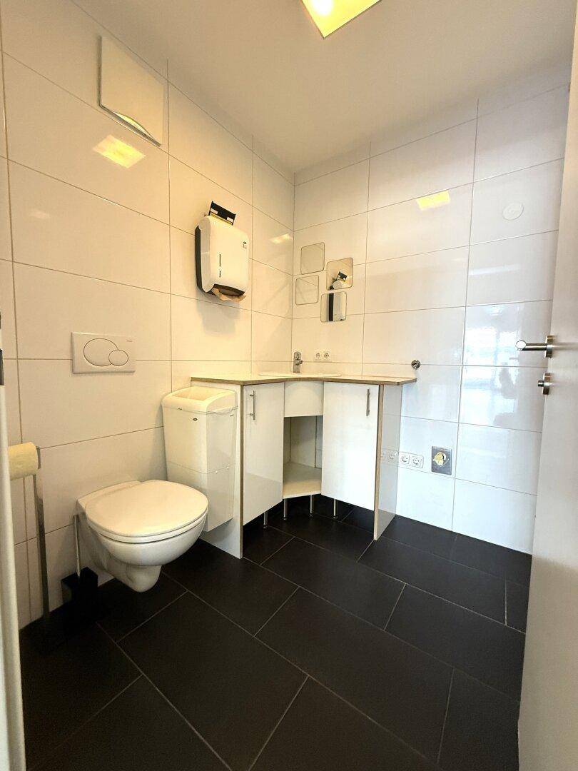 Modern geflieste Toilette mit Wand-WC, kleinem Waschbecken und dunklem Boden in sauberem Zustand.