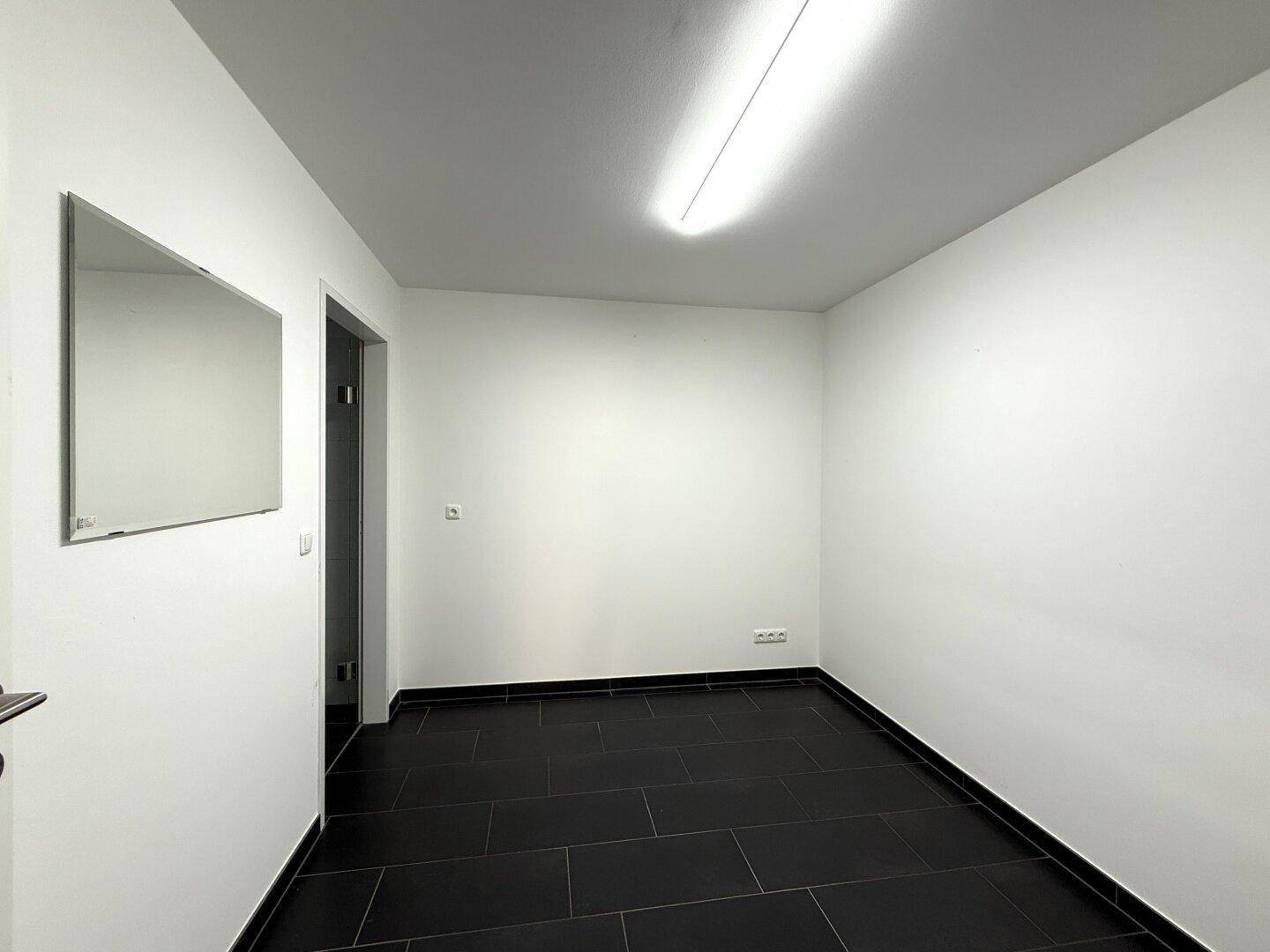 Schmaler Raum mit dunklen Fliesen, weißen Wänden und einem Spiegel an der Wand, ideal als Durchgang.