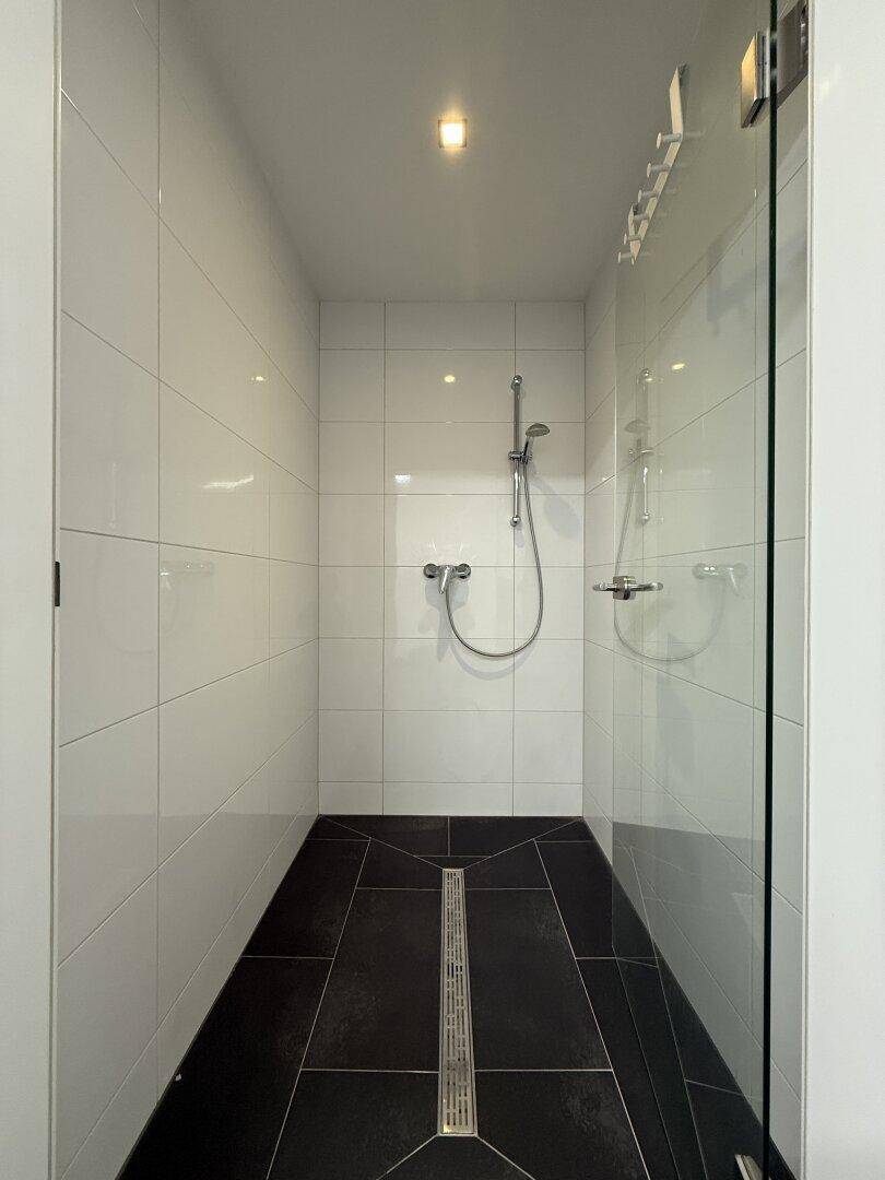 Moderne Duschkabine mit Glaswand, weißen Fliesen und dunklem Boden mit Ablaufrinne.