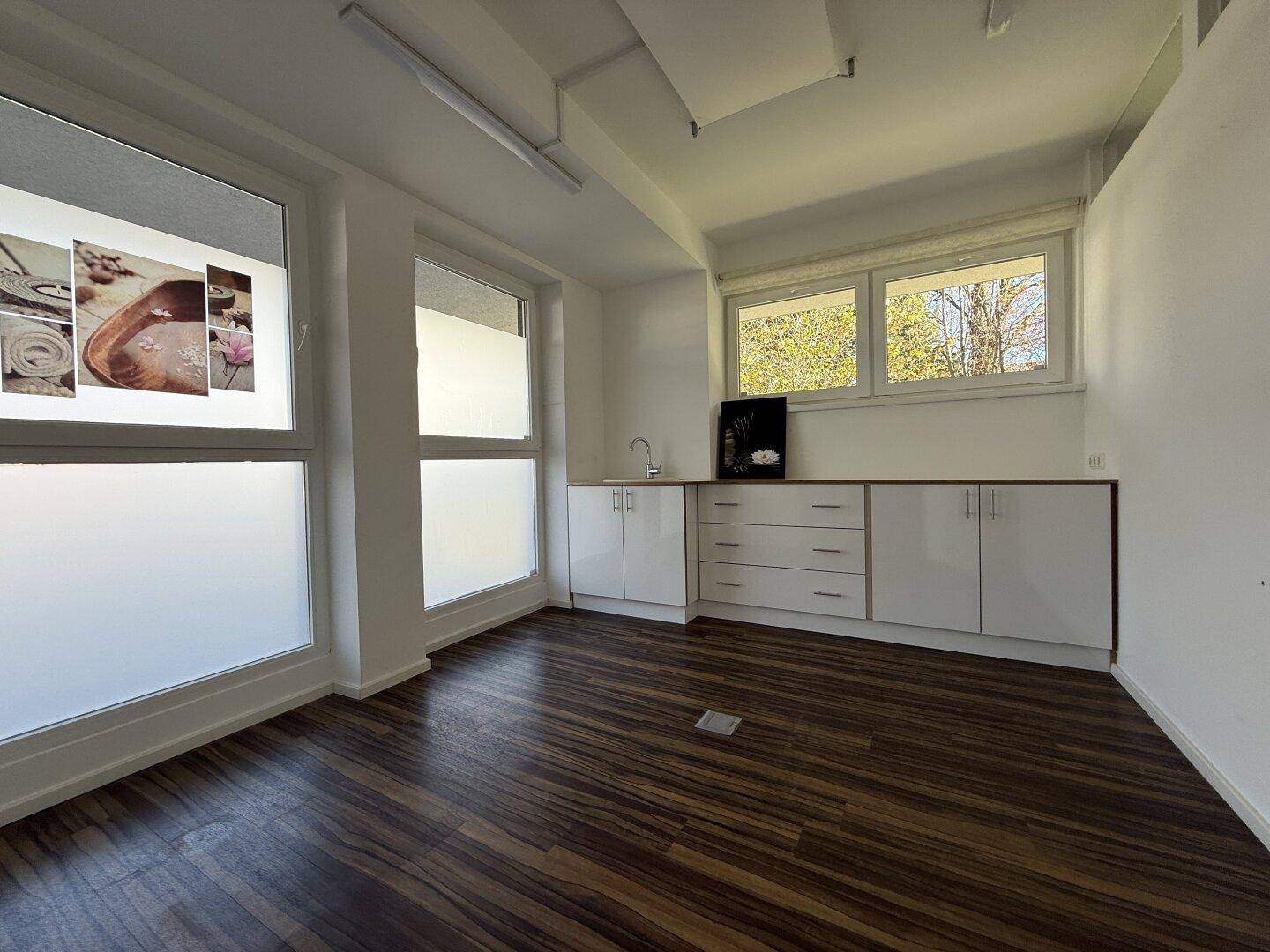 Helles Büro mit mehreren Fenstern, Laminatboden und einer einfachen Küchenzeile für den täglichen Gebrauch.