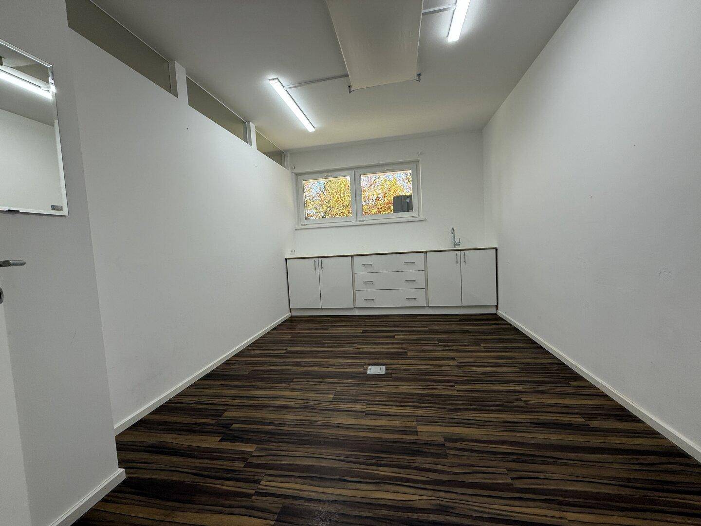 Geräumiges Büro mit Laminatboden, weißen Wänden und einer praktischen Küchenzeile unter den Fenstern.
