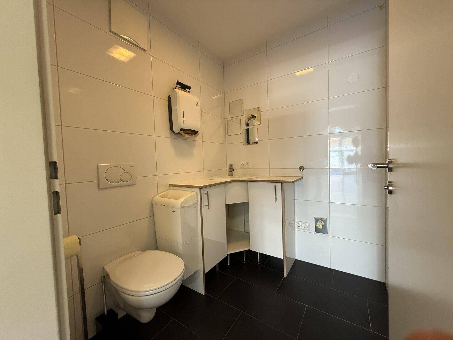 Modern geflieste Toilette mit Wand-WC, kleinem Waschbecken und dunklem Boden in sauberem Zustand.