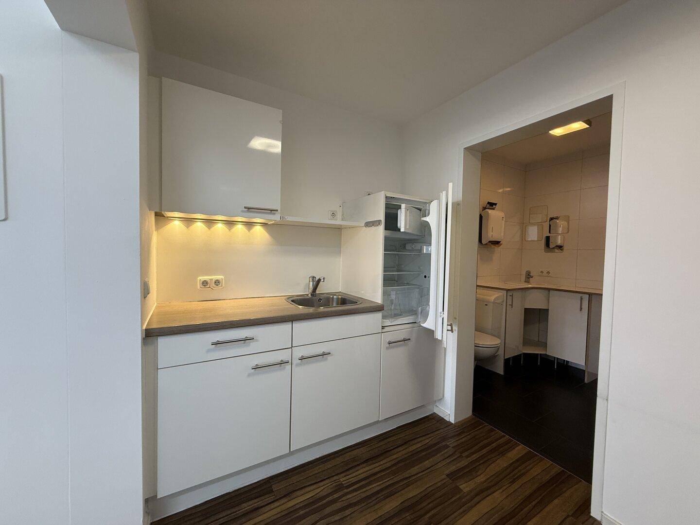 Kompakte Küchenzeile mit Spüle, Kühlschrank und weißen Schränken, angrenzend an einen Flur.