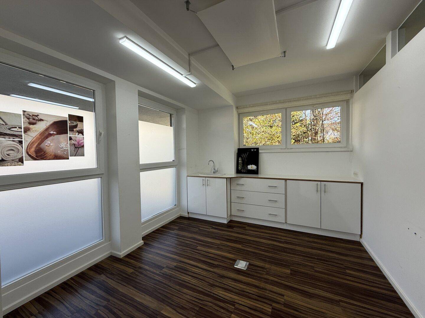 Helles Büro mit mehreren Fenstern, Laminatboden und einer einfachen Küchenzeile für den täglichen Gebrauch.
