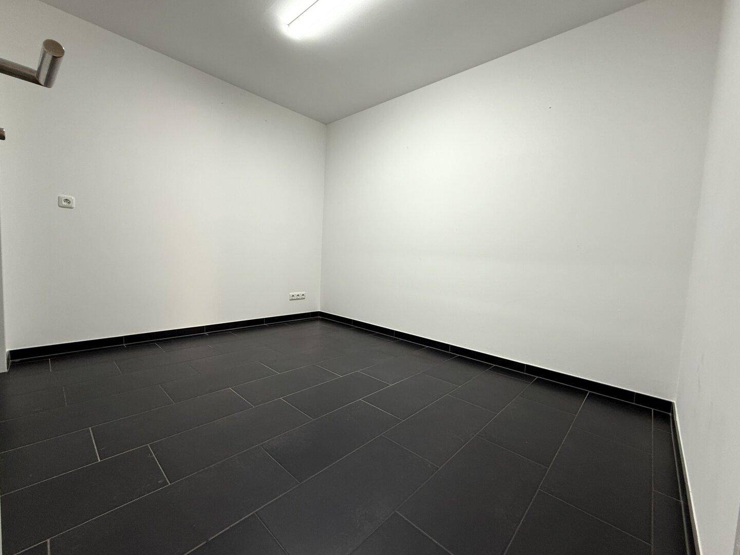 Kleiner, leerer Raum mit dunklen Fliesen und weißen Wänden, ideal als Abstellraum.