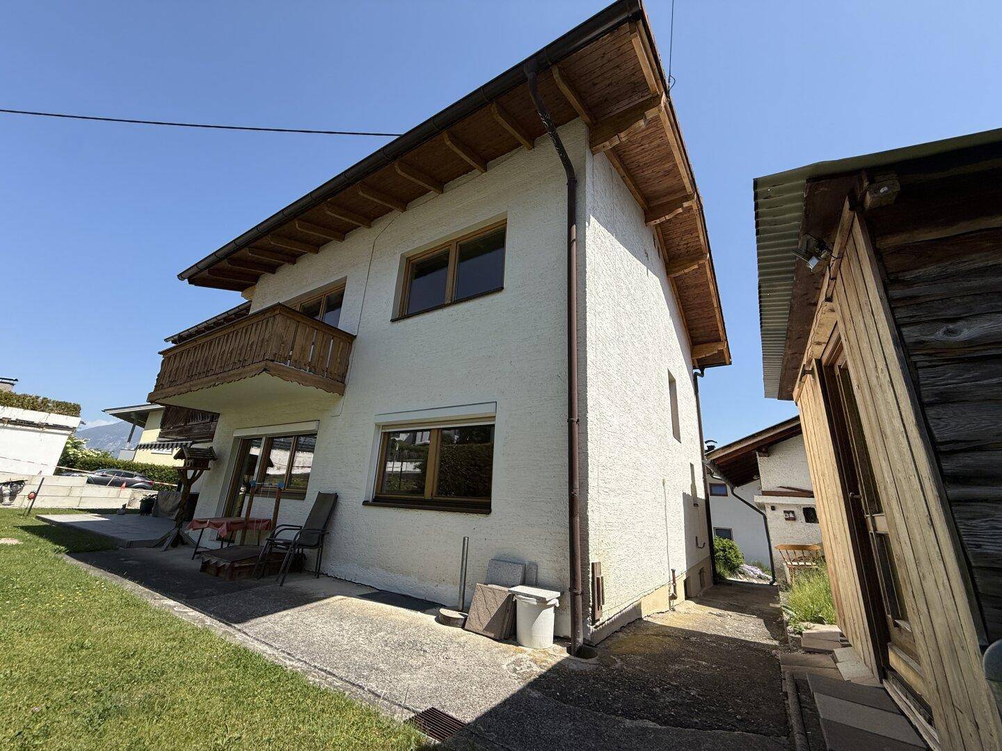 Seitenansicht des Hauses mit kleiner Terrasse, Gartenbereich und Holzbalkon im Obergeschoss.