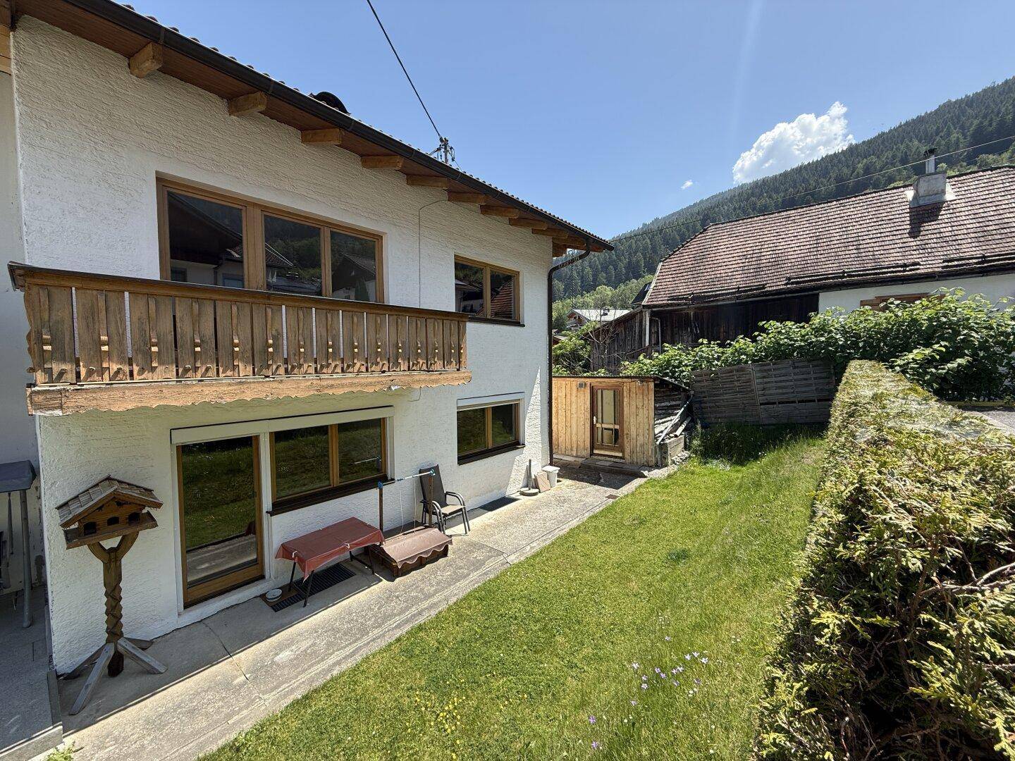 Hausfassade mit grünem Garten, kleiner Terrasse und Blick auf die umliegenden Berge.