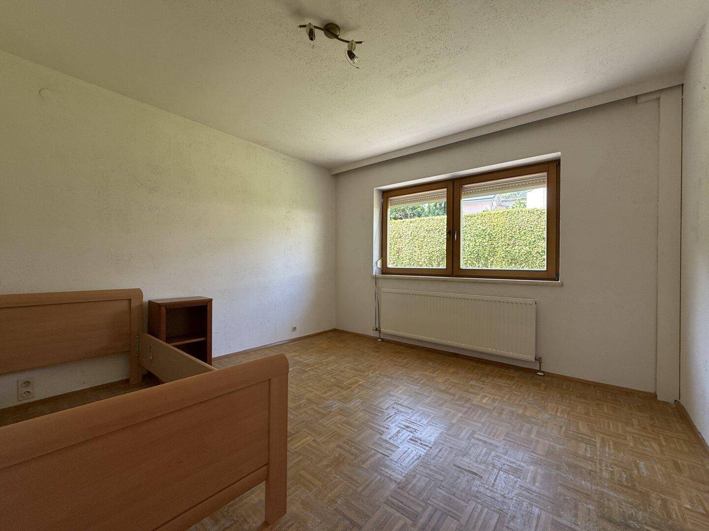 Schlafzimmer mit Parkettboden, großem Fenster und Blick auf die grüne Hecke.