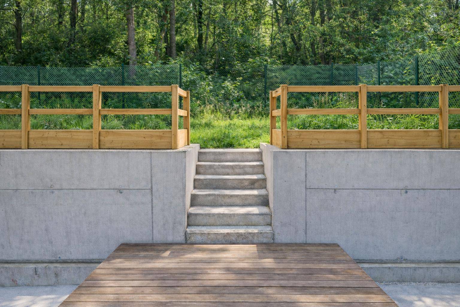 Holzterrasse mit Betontreppe, die zu einem bewaldeten Bereich mit Holzzaun führt.