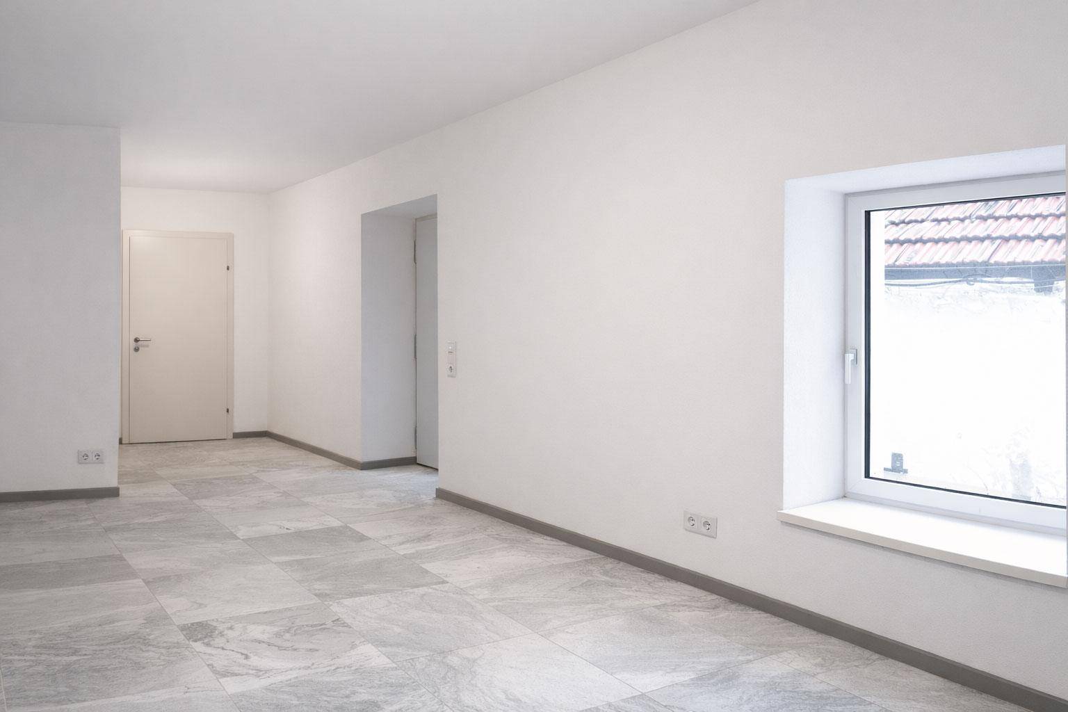 Leerer, heller Flur mit großen Fliesen und einem Fenster, das Tageslicht hereinlässt.