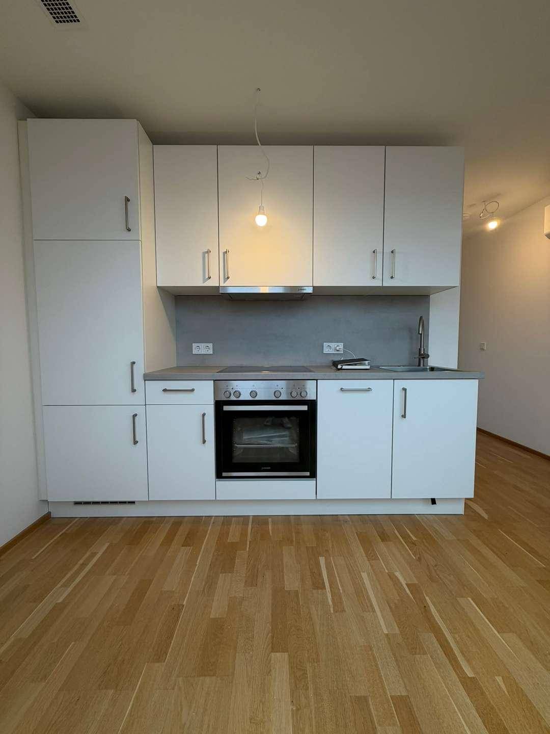 Kompakte Einbauküche mit weißen Schränken, Herd und Spüle in offenem Raum.
