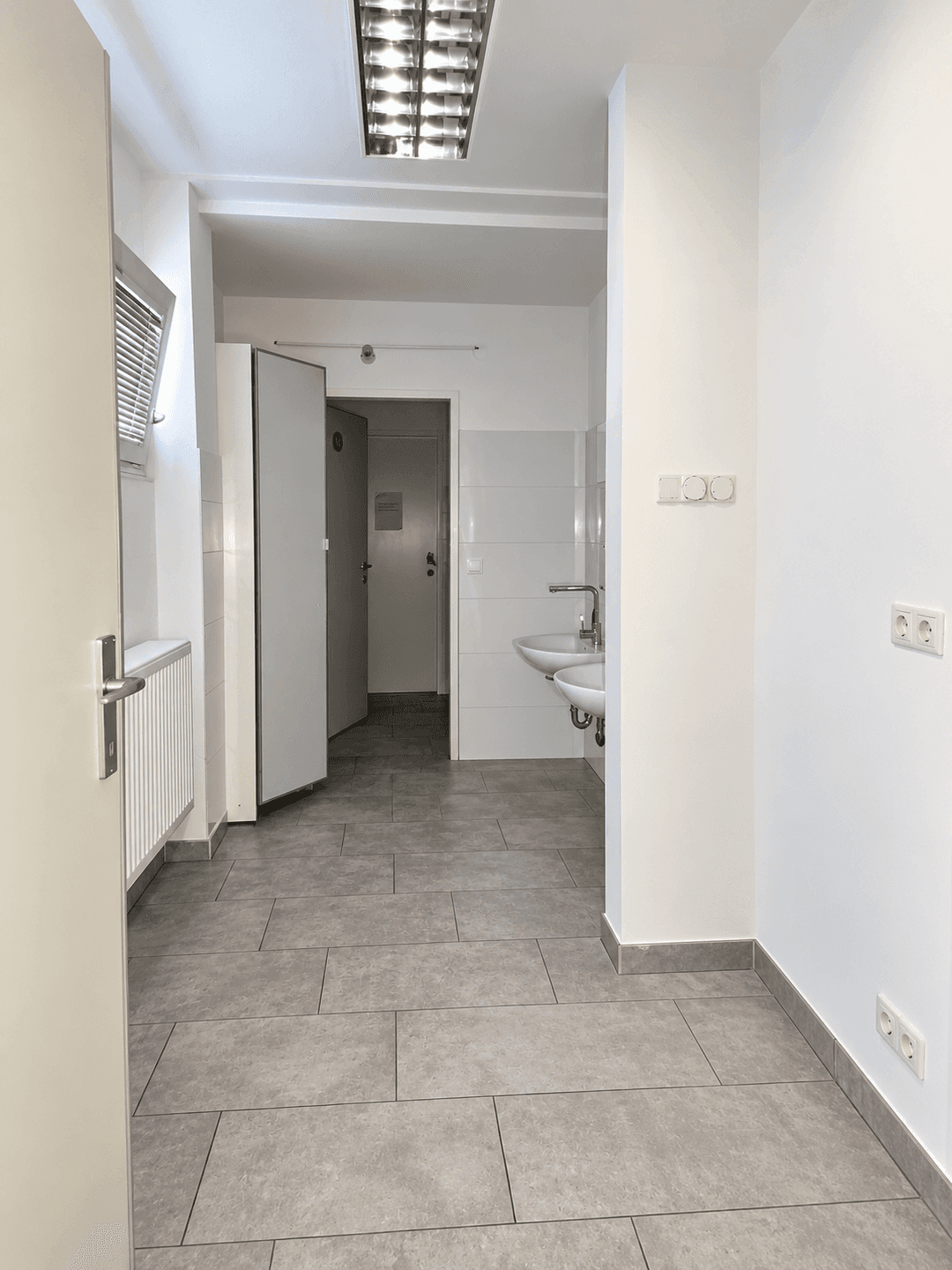 Heller Sanitärbereich mit Fliesenboden, Waschbecken und Zugang zu weiteren Räumen.