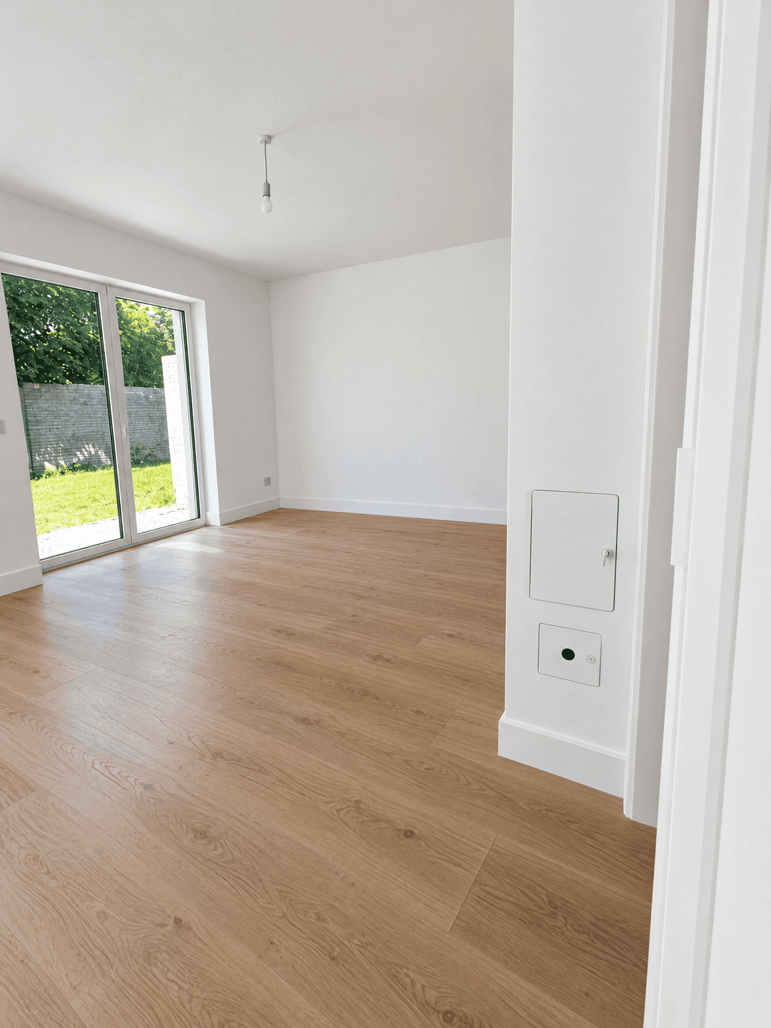 Helles, leeres Zimmer mit Holzboden und großen Glastüren, die zum Garten führen.