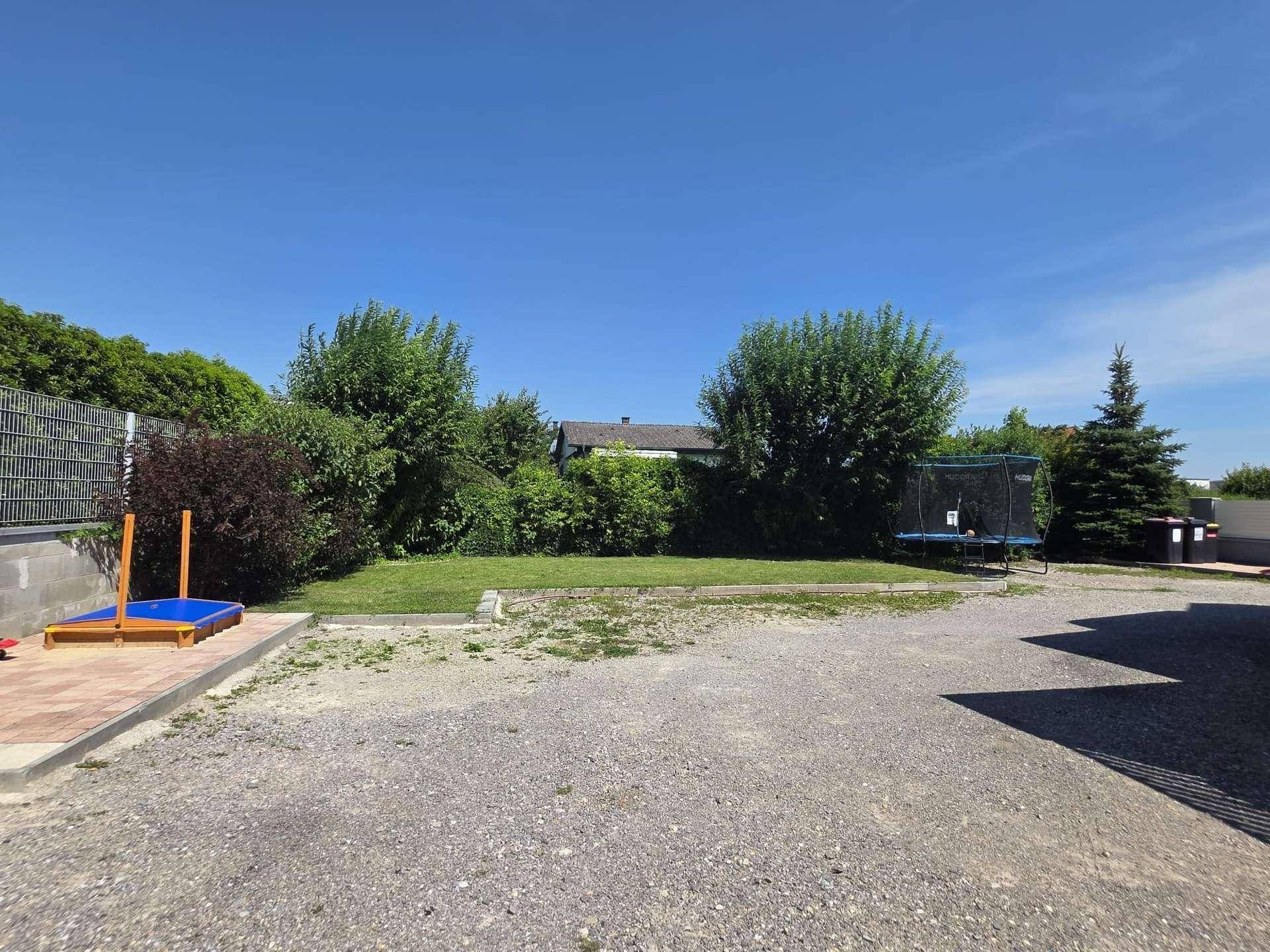Großer Garten mit Rasenfläche, Sandkasten und Trampolin unter blauem Himmel.