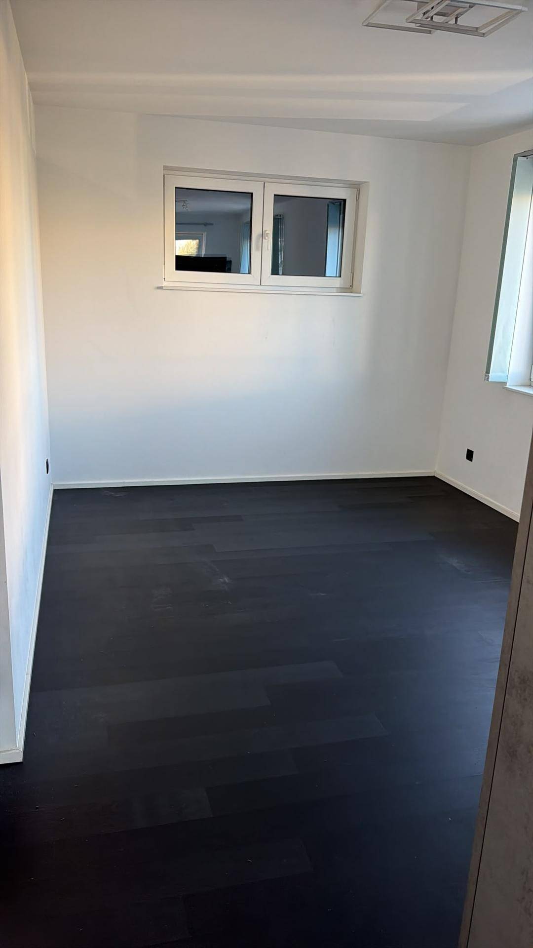 Leerer, heller Raum mit dunklem Holzboden und einem Fenster für natürliches Licht.