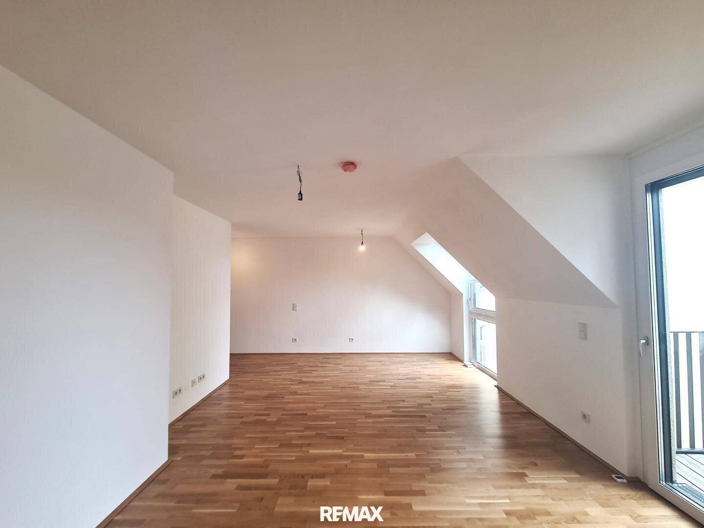 Geräumiger, heller Raum mit Holzboden, weißen Wänden und Zugang zu einem Balkon oder einer Terrasse.
