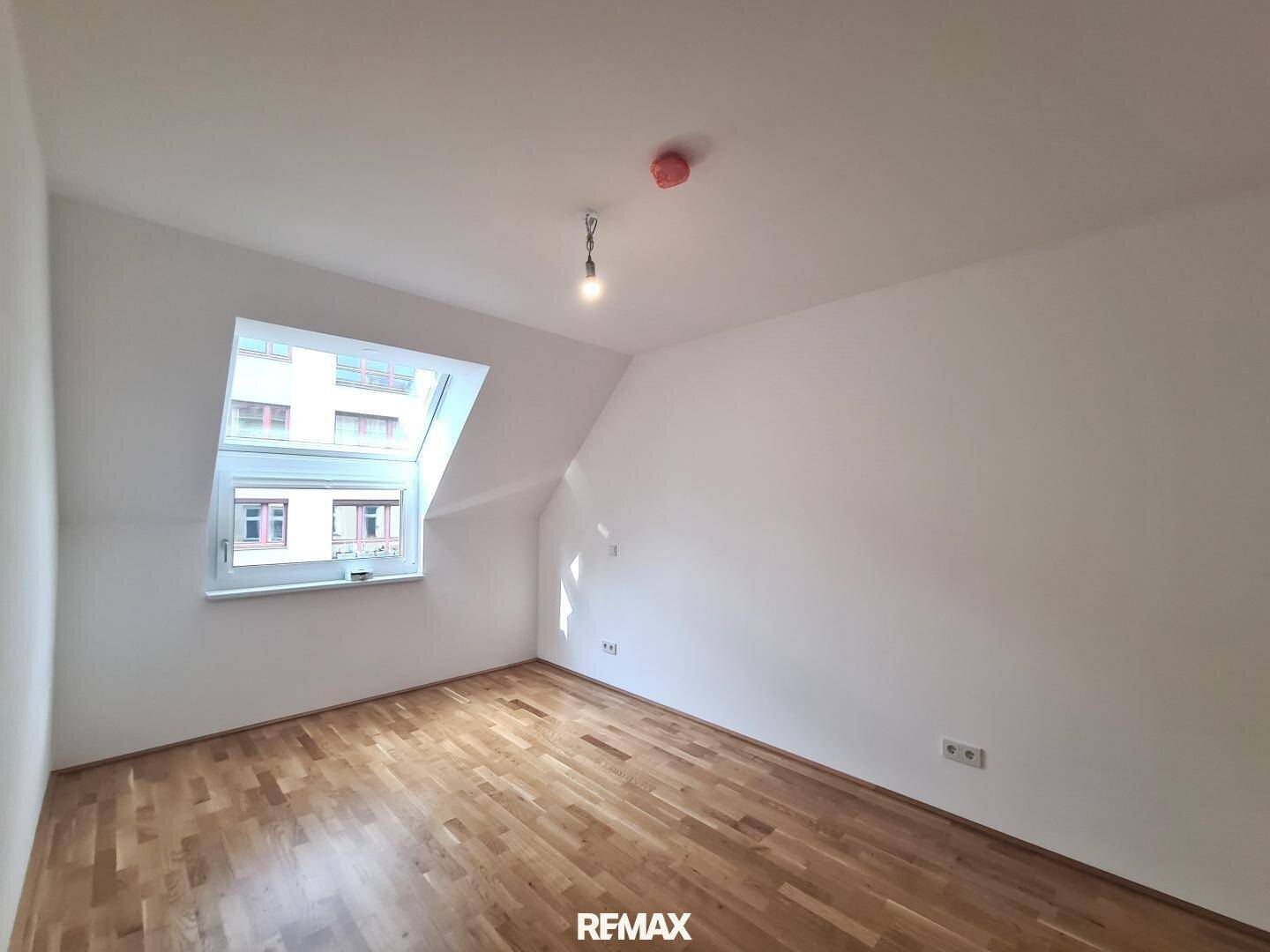 Helles Schlafzimmer mit Holzboden, weißen Wänden und einem Dachfenster, das viel Tageslicht hereinlässt.