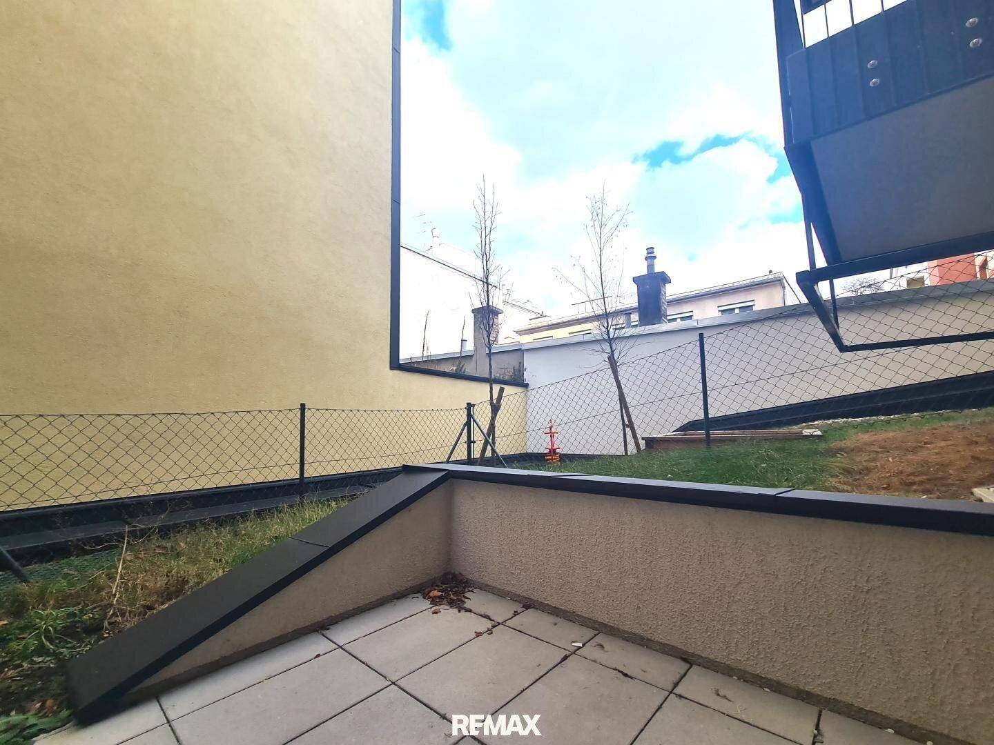Kleine, geflieste Terrasse im Freien, umgeben von einer niedrigen Mauer und Grünfläche.