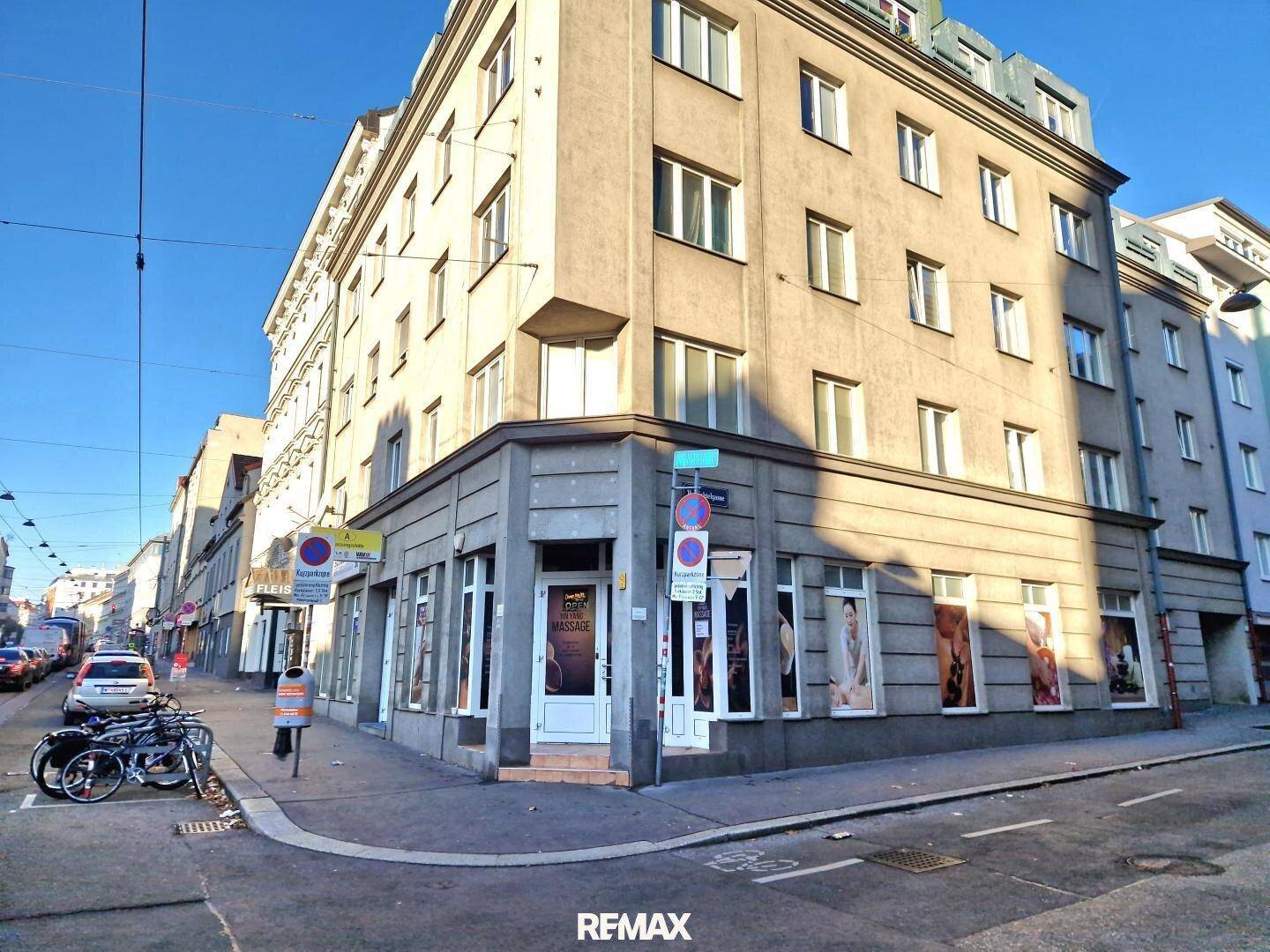 Eckgebäude an einer belebten Straße mit Geschäften im Erdgeschoss unter blauem Himmel.