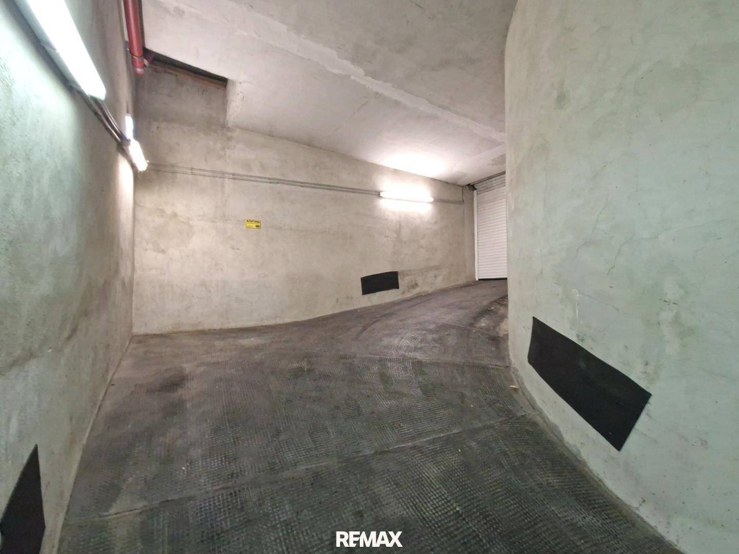 Kurvige Zufahrtsrampe in der Tiefgarage mit Betonwänden und einem Rolltor am Ende.