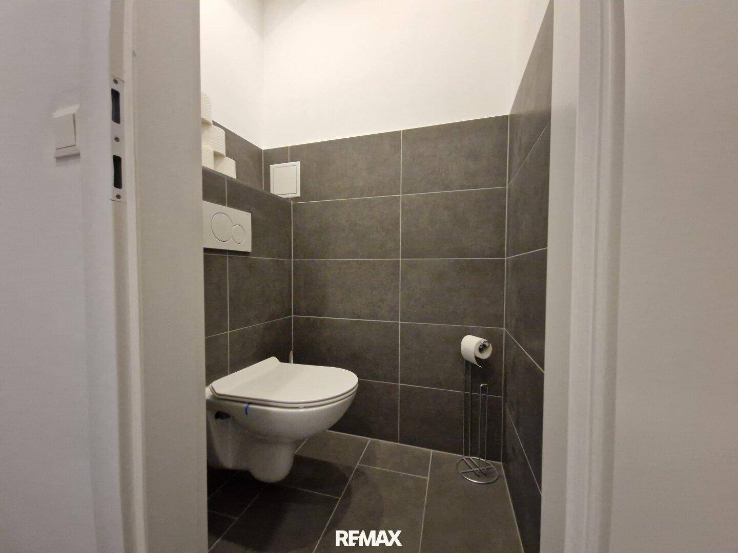 Separates WC mit modernen grauen Fliesen und wandhängender Toilette.