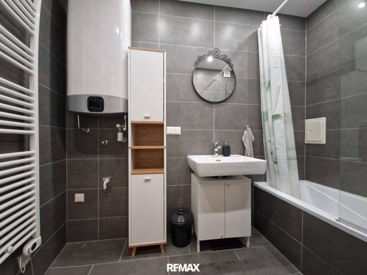 Modernes Badezimmer mit grauen Fliesen, Badewanne, Waschbecken und Handtuchheizkörper.