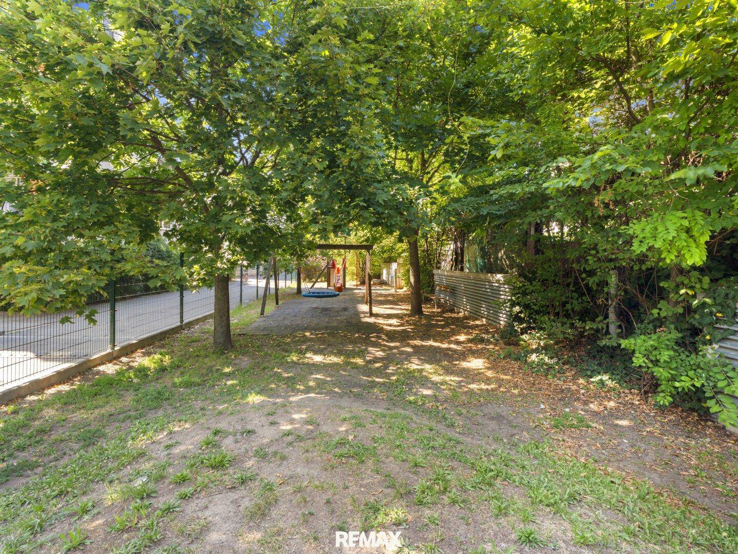 Spielplatzbereich im Freien mit Bäumen und Schaukeln, umgeben von einem Zaun.