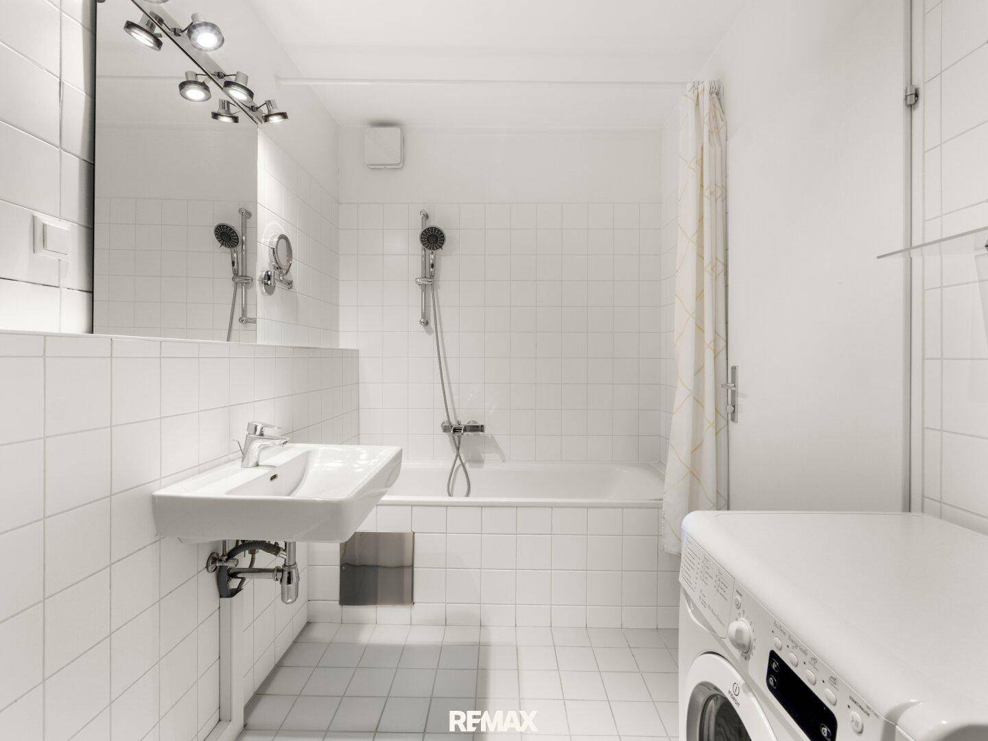 Helles Badezimmer mit weißen Fliesen, Badewanne mit Duschfunktion und integrierter Waschmaschine.
