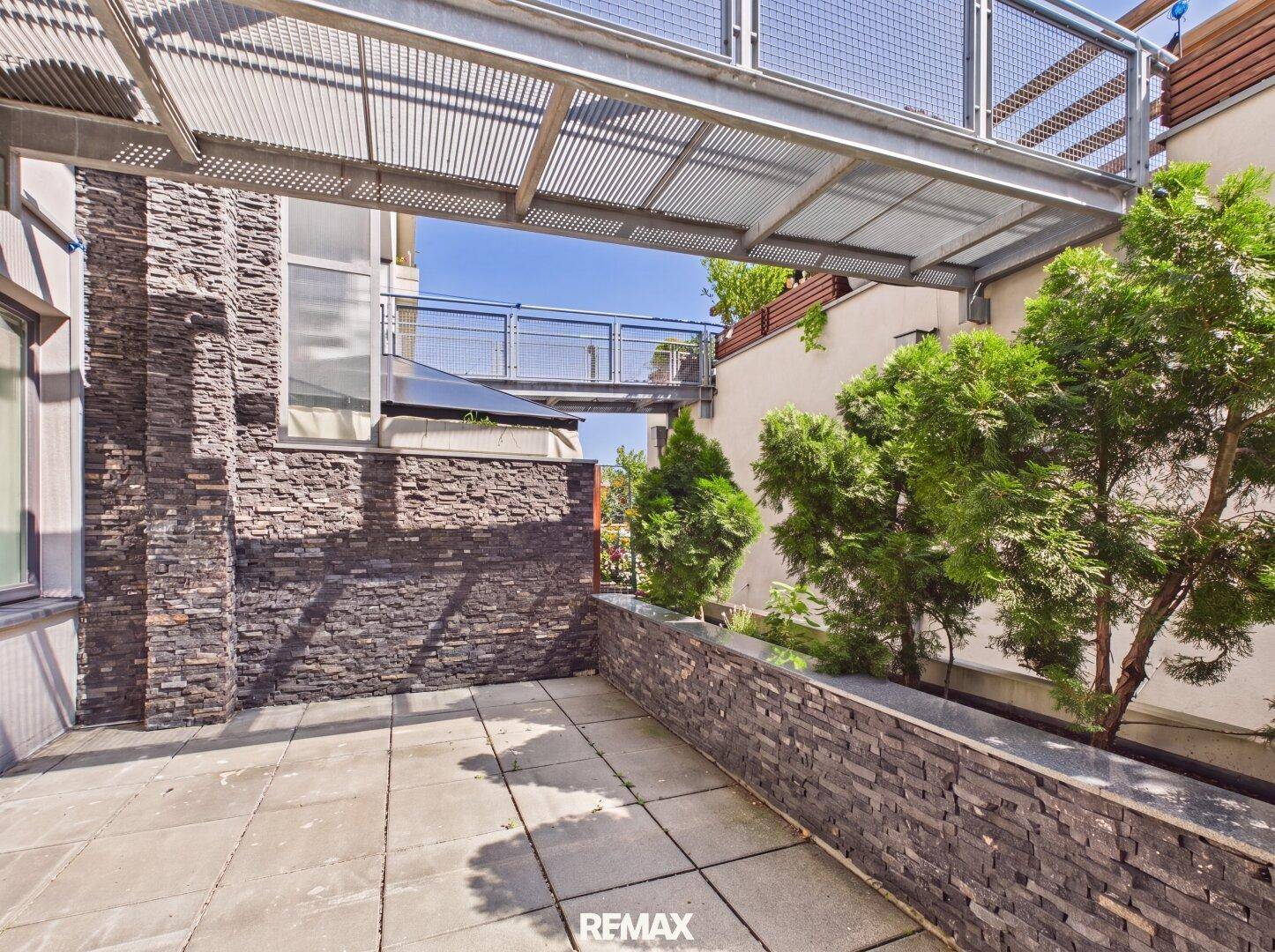 Gepflasterte Terrasse mit Steinmauern und Pflanzen, verbunden durch eine moderne Metallbrücke.