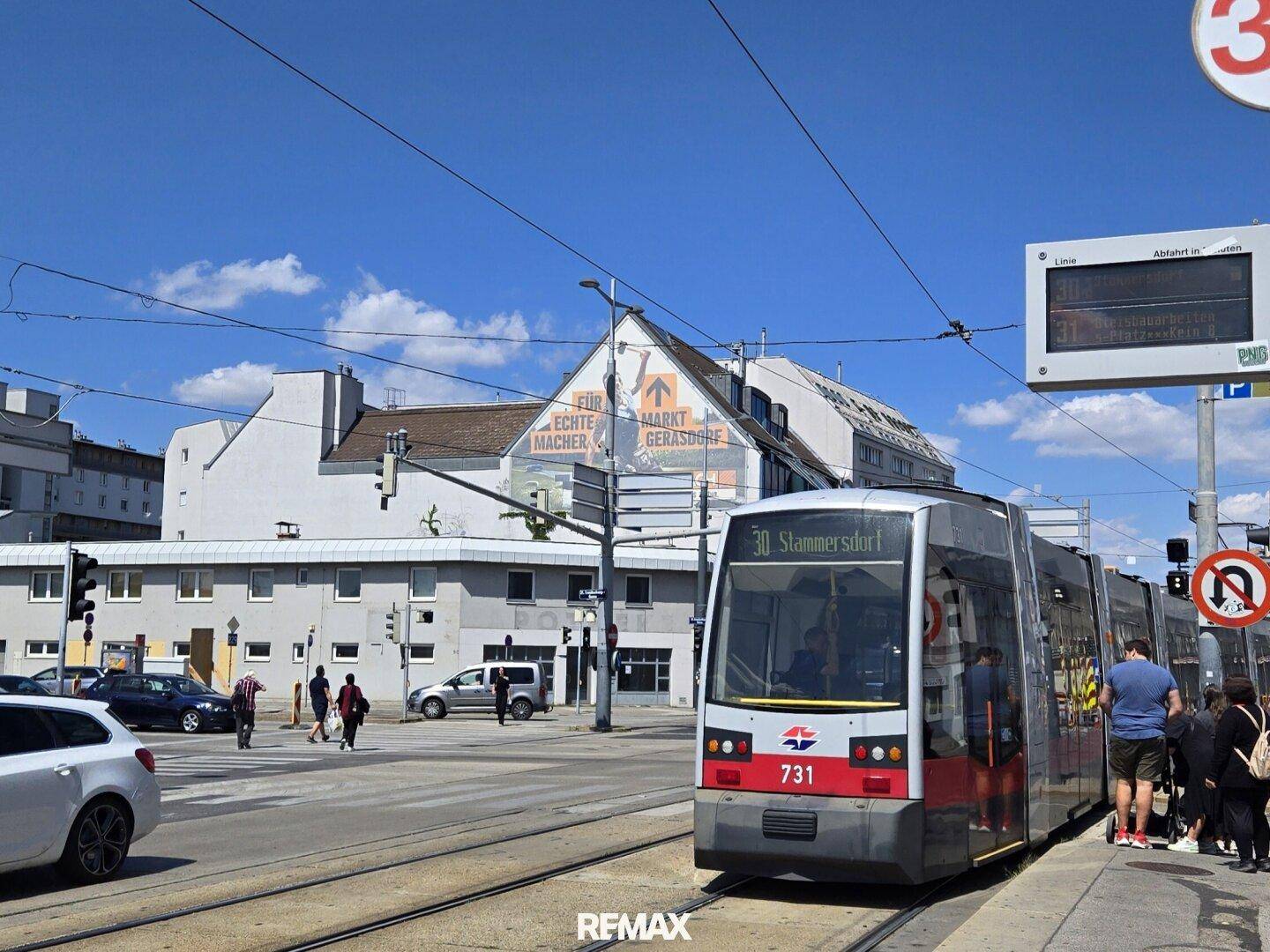 Straßenansicht mit einer Straßenbahn, Passanten und Gebäuden unter blauem Himmel in der Stadt.