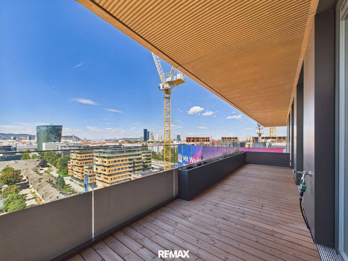 Geräumiger Balkon mit Holzboden und Blick auf die Stadt und eine Baustelle mit Kran.