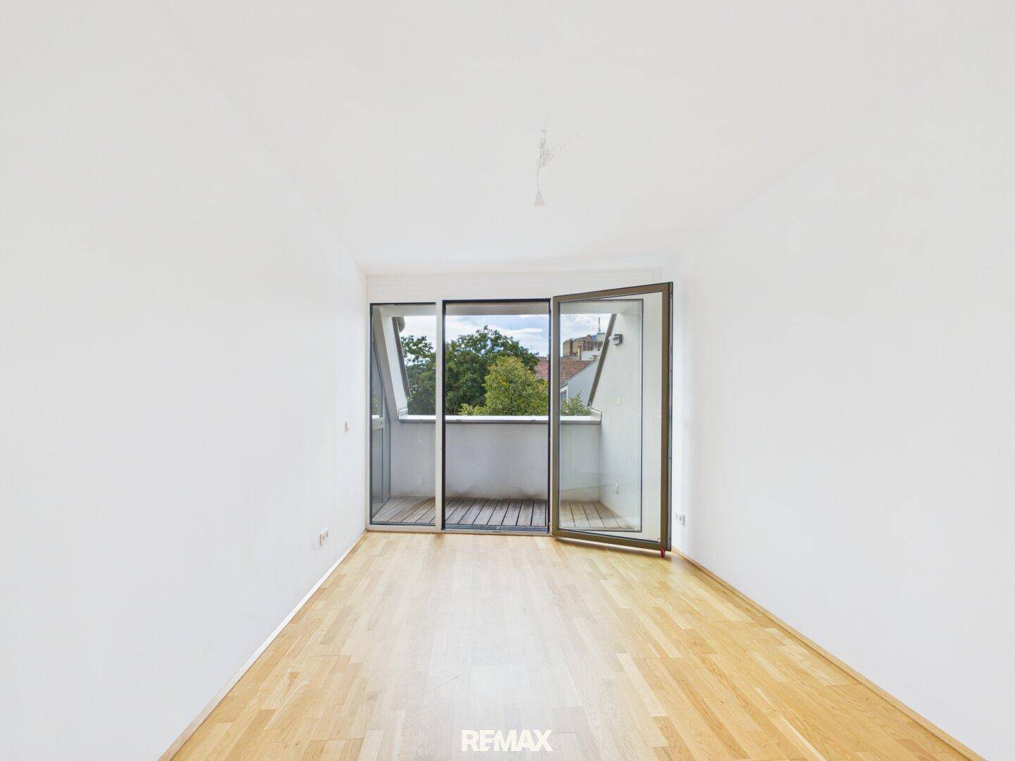 Leeres Schlafzimmer mit hellem Parkettboden und Zugang zu einem kleinen Balkon mit grüner Aussicht.